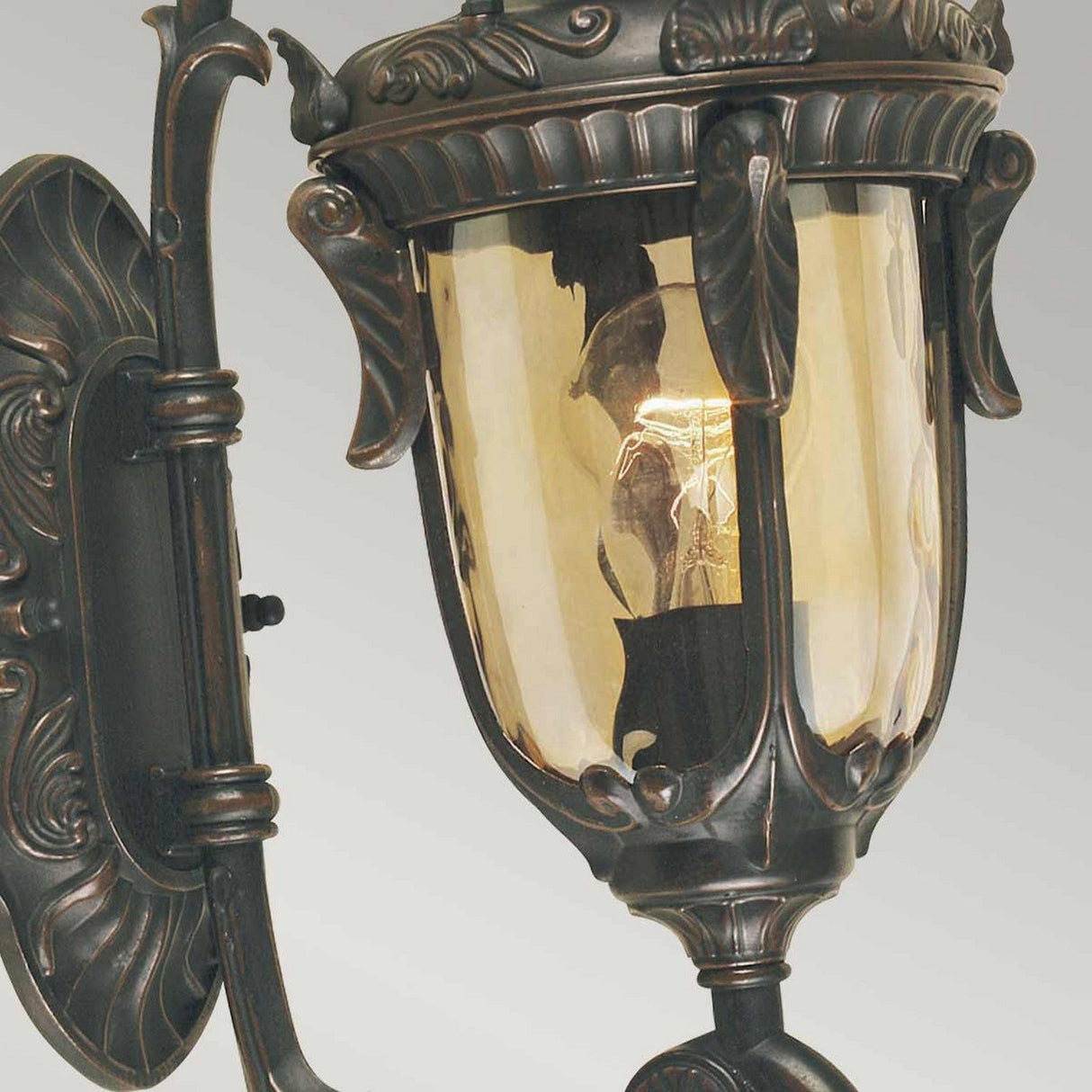 A close-up of the Philadelphia 1 Light Outdoor Small Wall Light reveals its decorative metal frame and glass panels. This lantern, with period-inspired designs, encases a glowing light bulb. The metal's old bronze patina finish beautifully enhances its intricate floral patterns.