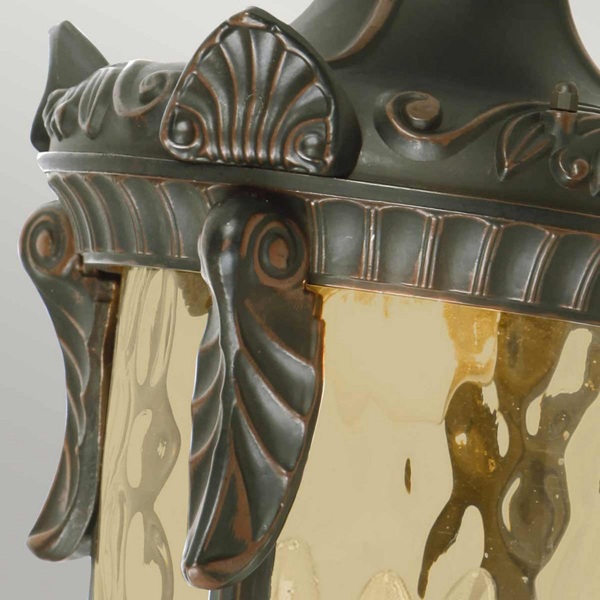 Close-up of the Philadelphia 3 Light Outdoor Large Down Wall Light in Old Bronze, featuring a textured glass shade. The intricately designed bronze frame highlights elegant scrollwork and decorative shell motifs, offering period charm. The warm amber-tinted glass enhances its antique design aesthetic.