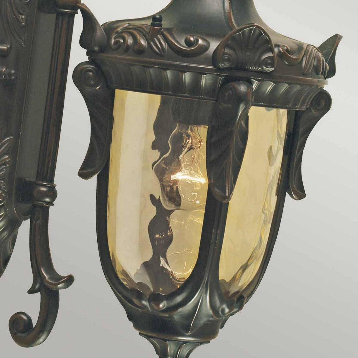 A close-up of the Philadelphia 1 Light Outdoor Medium Down Wall Light in Old Bronze showcases its ornate design, featuring yellow-tinted glass and intricate metalwork with leaf and scroll motifs. The glowing bulb inside highlights its vintage-style exterior lighting.