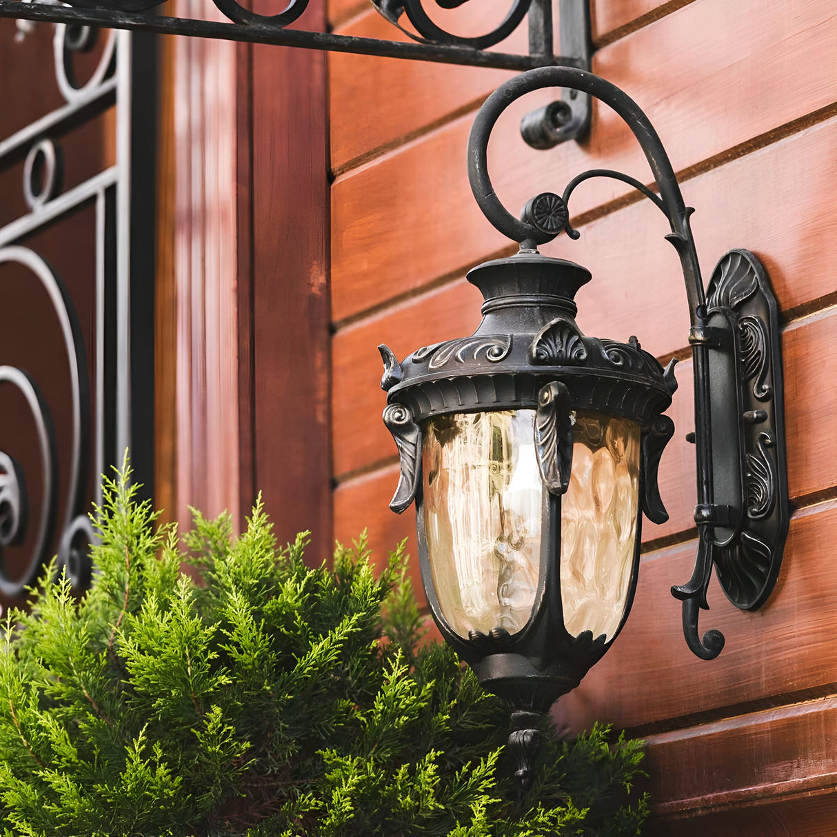 The Philadelphia 1 Light Outdoor Medium Down Wall Light in Old Bronze boasts a decorative black wrought iron lantern design with frosted glass. Mounted on a wooden wall and surrounded by green shrubbery, it brings charm and elegance to the setting, while its ornate metal design accentuates the exterior lighting aesthetic in the background.