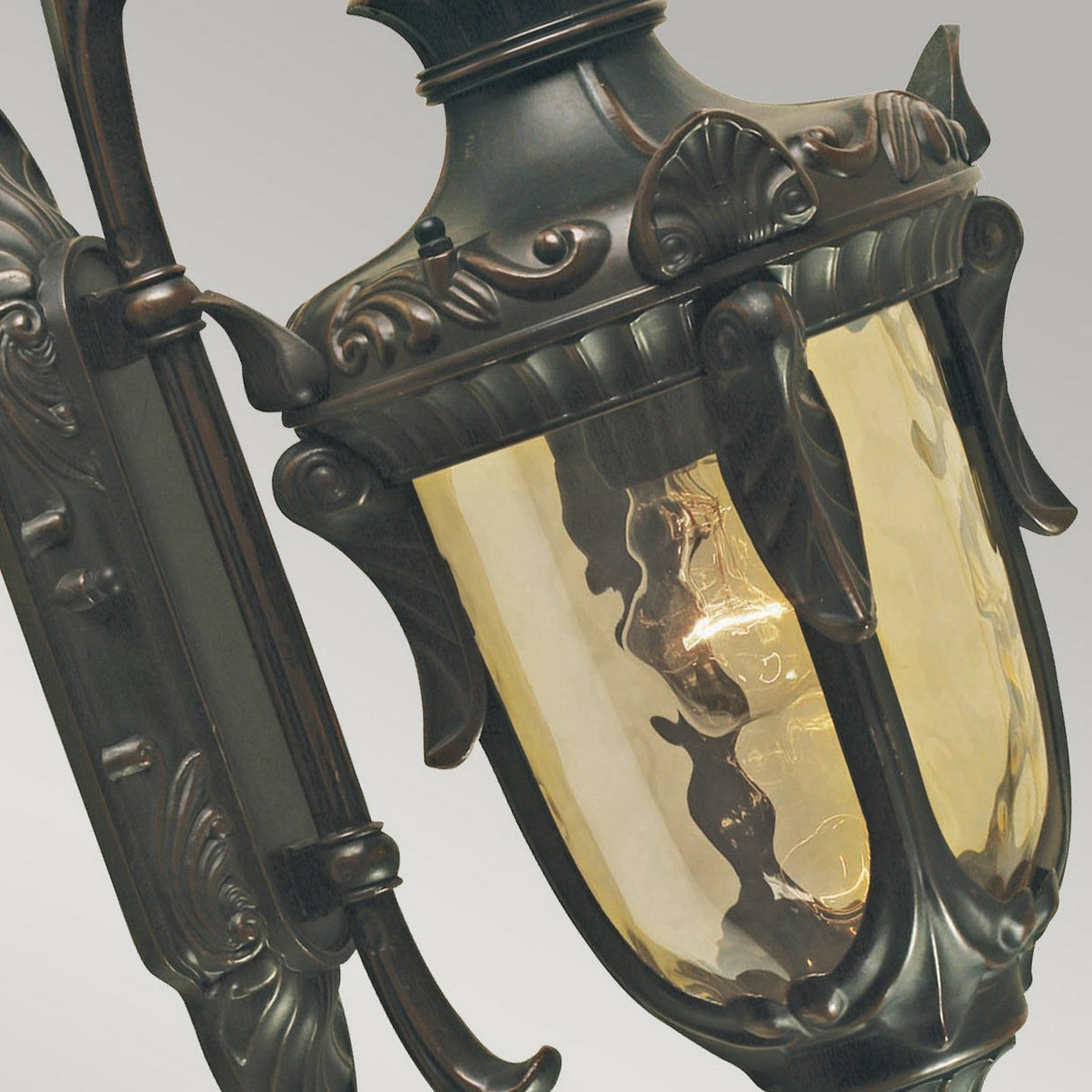A close-up view of the Philadelphia 1 Light Outdoor Small Down Wall Light showcases its ornate, antique-style design with an old bronze patina finish. The glowing bulb inside and slightly tinted glass enhance the vintage appeal of this charming fixture.