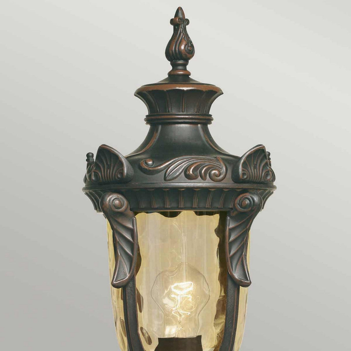 The Philadelphia 1 Light Medium Pedestal in Old Bronze is designed with an ornate, vintage-style featuring intricate dark metal detailing and a decorative top. Its champagne-tinted glass showcases a visible bulb inside. Ideal for enhancing any outdoor lighting setup with charm, it stands elegantly against a light gray background.