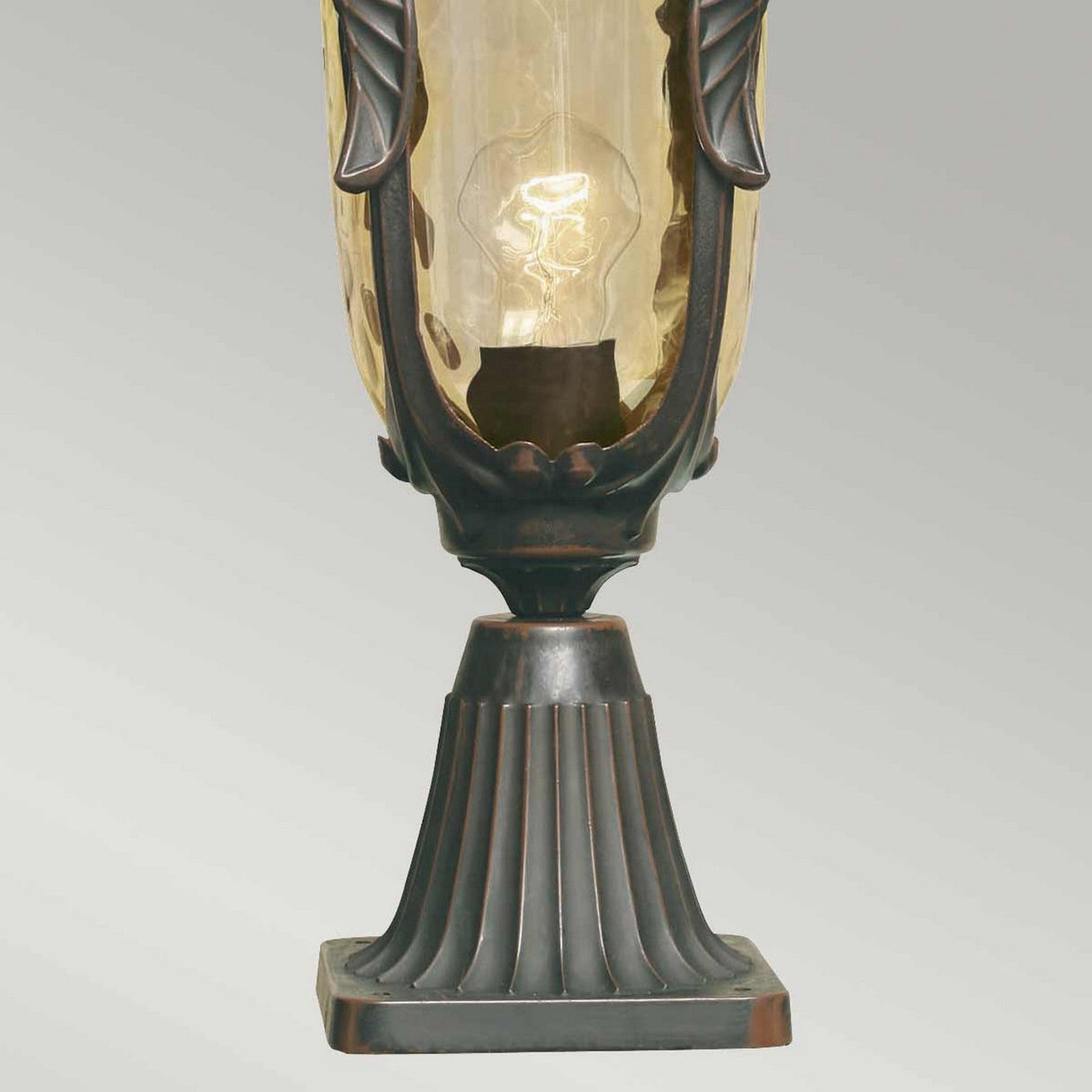 A close-up of the Philadelphia 1 Light Medium Pedestal in Old Bronze, featuring a decorative amber glass shade that beautifully showcases an illuminated bulb inside. The champagne-tinted glass enhances its elegance, while the neutral grey background highlights its timeless charm.