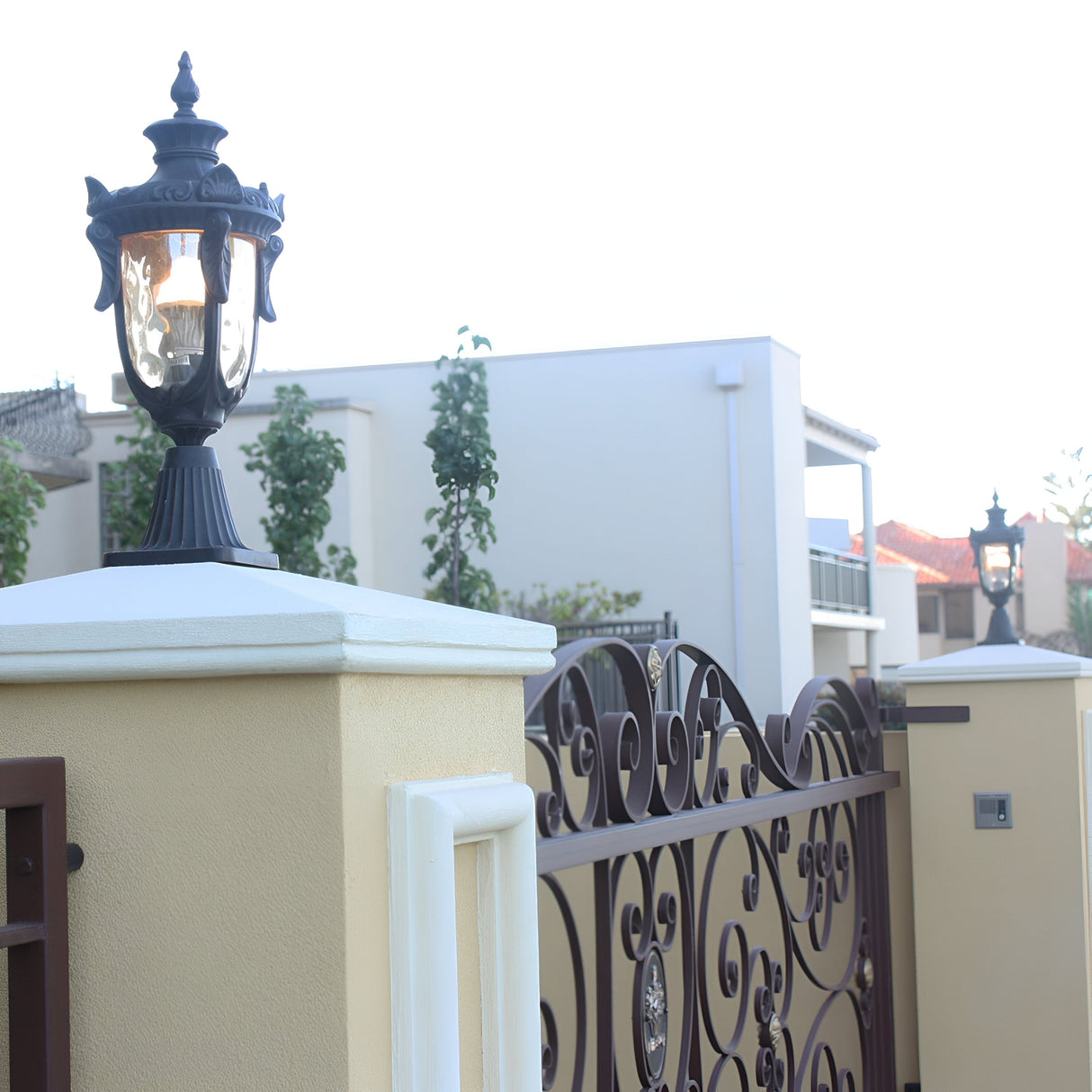 A Philadelphia 1 Light Medium Pedestal in Old Bronze, featuring champagne-tinted glass, stands on a cream-colored pillar beside an intricate wrought iron gate. In the background, a modern building with a flat roof is framed by trees and a light blue sky, enhanced by elegant outdoor lighting.