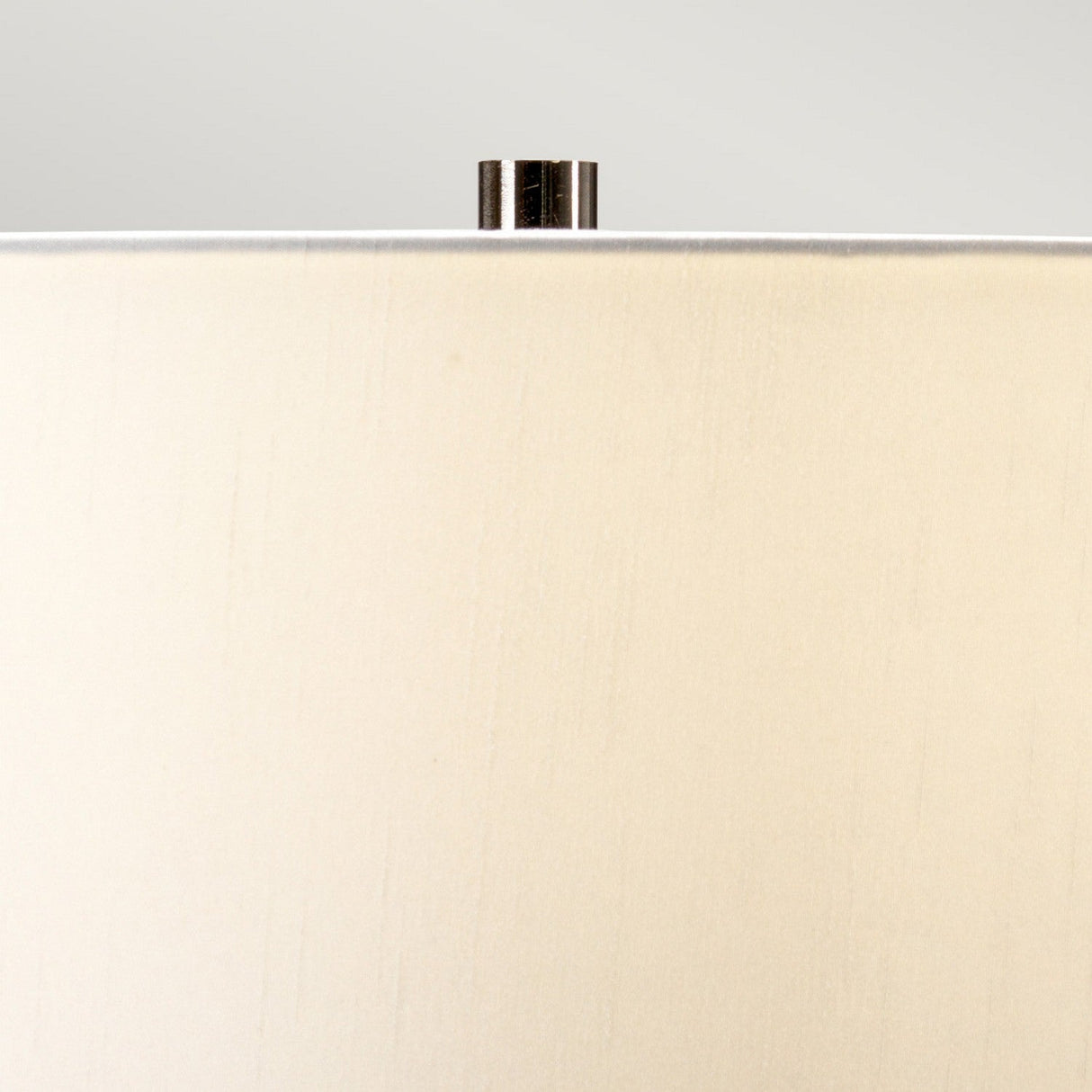 Close-up of a Pinner 1 Light Table Lamp in Subtle Pink and Matt Cream, highlighting the cream faux silk cylinder shade adorned with a metallic finial. The image focuses on the upper portion against a neutral background, drawing attention to the texture and color of the fabric.