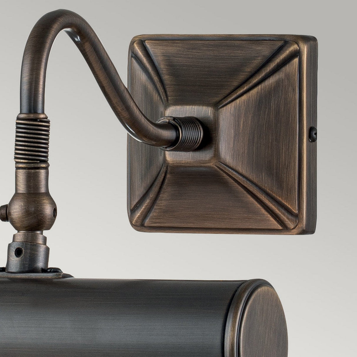 A close-up of the Picture Light 1 Light Small - Dark Bronze reveals its vintage-style design with a decorative square mounting plate and an adjustable, curved arm. The dark bronze finish provides an antique appearance, and the fixture boasts a cylindrical housing. The neutral gray wall enhances its elegant design reminiscent of a classic picture light.