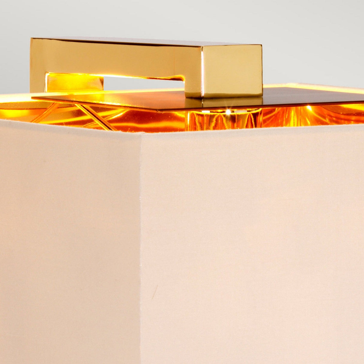 Close-up of the Plein 1 Light Table Lamp in Dark Brown/Polished Gold, showcasing its square beige shade and polished gold handle on top. The light inside emits a warm glow, softly illuminating through the shade. The backdrop is a plain, neutral color, accentuating the modern decor vibe.