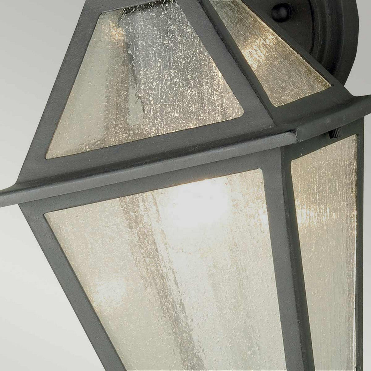 Close-up of the Polruan 1 Light Outdoor Wall Light in black, showcasing its geometric design. This fixture is IP44 rated for durability and features seeded glass panels and a sleek black metal frame, providing a gentle illumination from within.