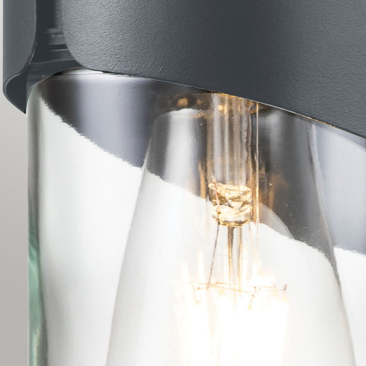Close-up of the Porto 1 Light Outdoor Wall Light in grey, showcasing a contemporary design with a transparent glass shade and visible bulb. Its upper section emulates a stylish nautical lantern made from smooth, dark gray material that elegantly curves over the glass, accentuating natural light reflections and detailed bulb filaments.