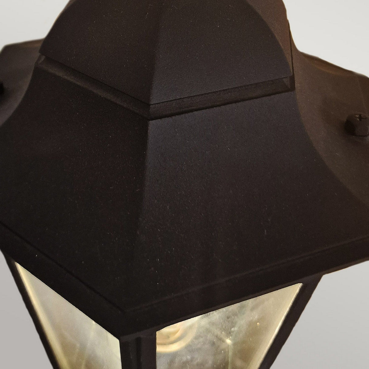 Close-up of the Parish 1 Light Outdoor Up Wall Light in a black finish, featuring a hexagonal design with a glass panel. The internal light emits a faint glow, creating a soft ambiance. This IP44-rated lantern is set against a plain, light gray background.