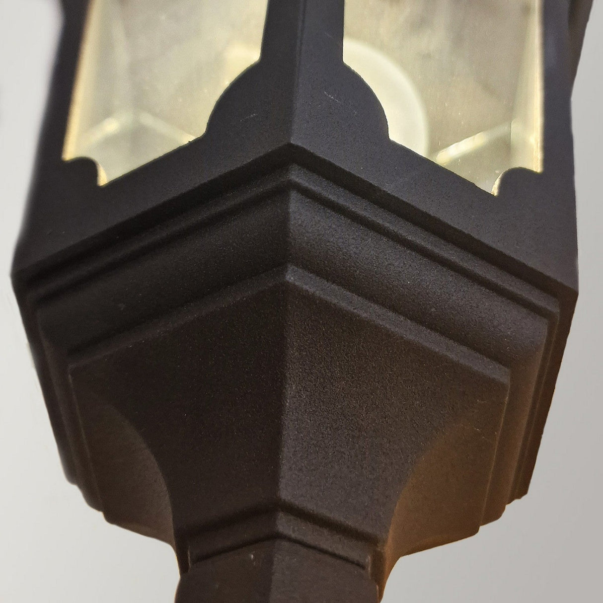 Close-up of the Parish 1 Light Outdoor Up Wall Light in black, showcasing its textured finish and IP44 rating. The lantern boasts a classic design with clear glass panes that highlight the light from a visible bulb inside. The angular base is prominently featured, emphasizing its geometric shape.