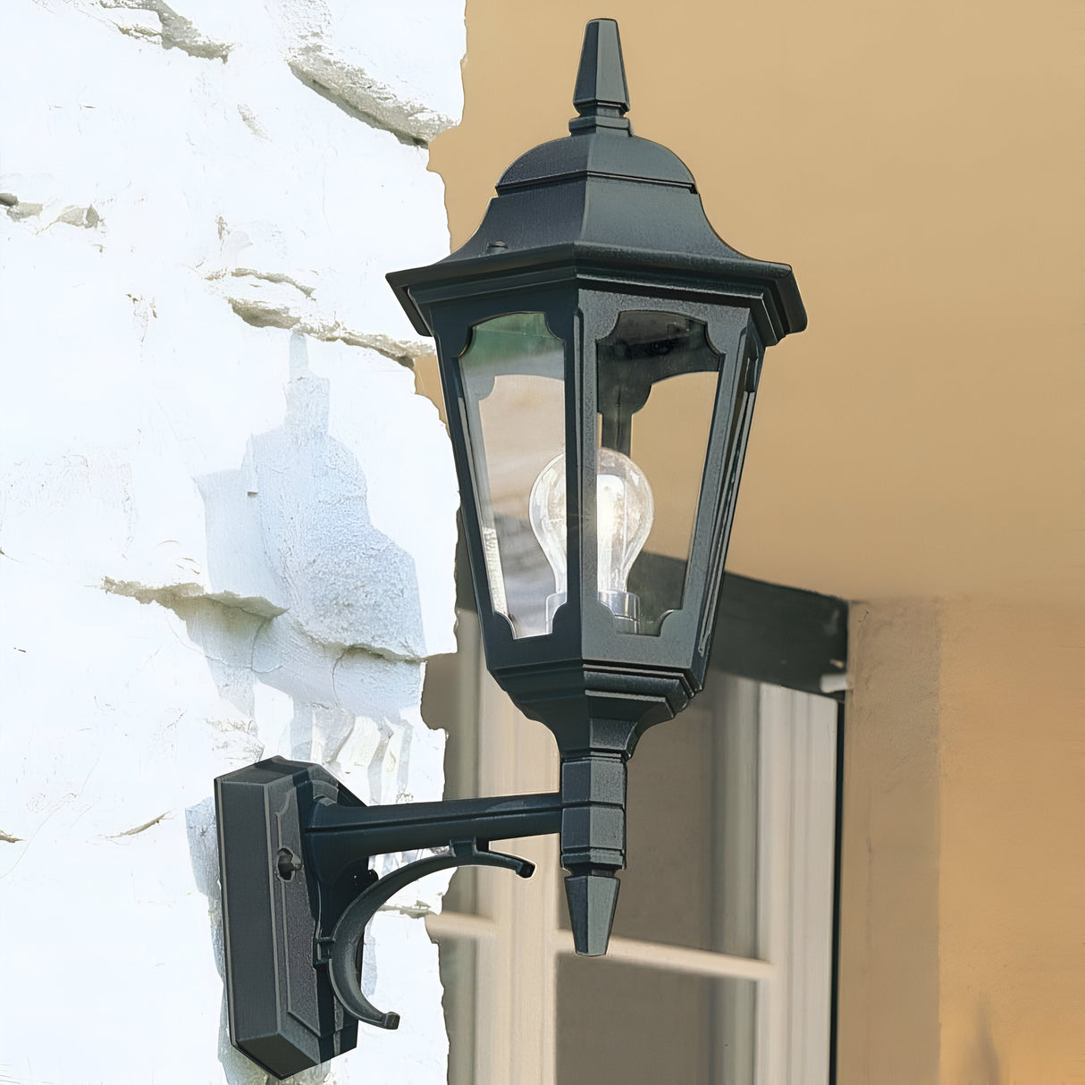 The Parish 1 Light Outdoor Up Wall Light in black finish is mounted on a white brick wall, with its glass-covered bulb casting a bright shadow. Featuring an IP44 rating for durability, the lantern perfectly complements the partially visible beige-framed window in the background.