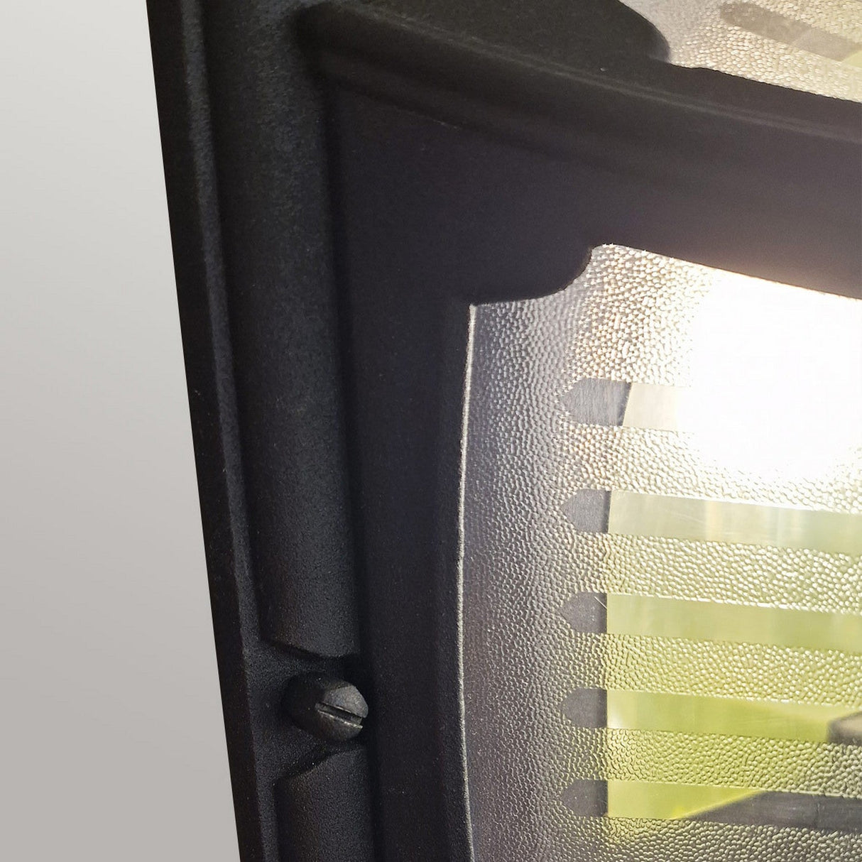 Close-up of the Parish 1 Light Outdoor Ceiling Flush - Black, showcasing its textured glass light fixture crafted with die-cast aluminum and enveloped in a sleek black metal frame. A screw is positioned near the bottom of the frame, enhancing its sturdy appearance, while the bright light casts striking horizontal lines across the glass. Ideal for outdoor use with its IP44 rating providing reliable durability.