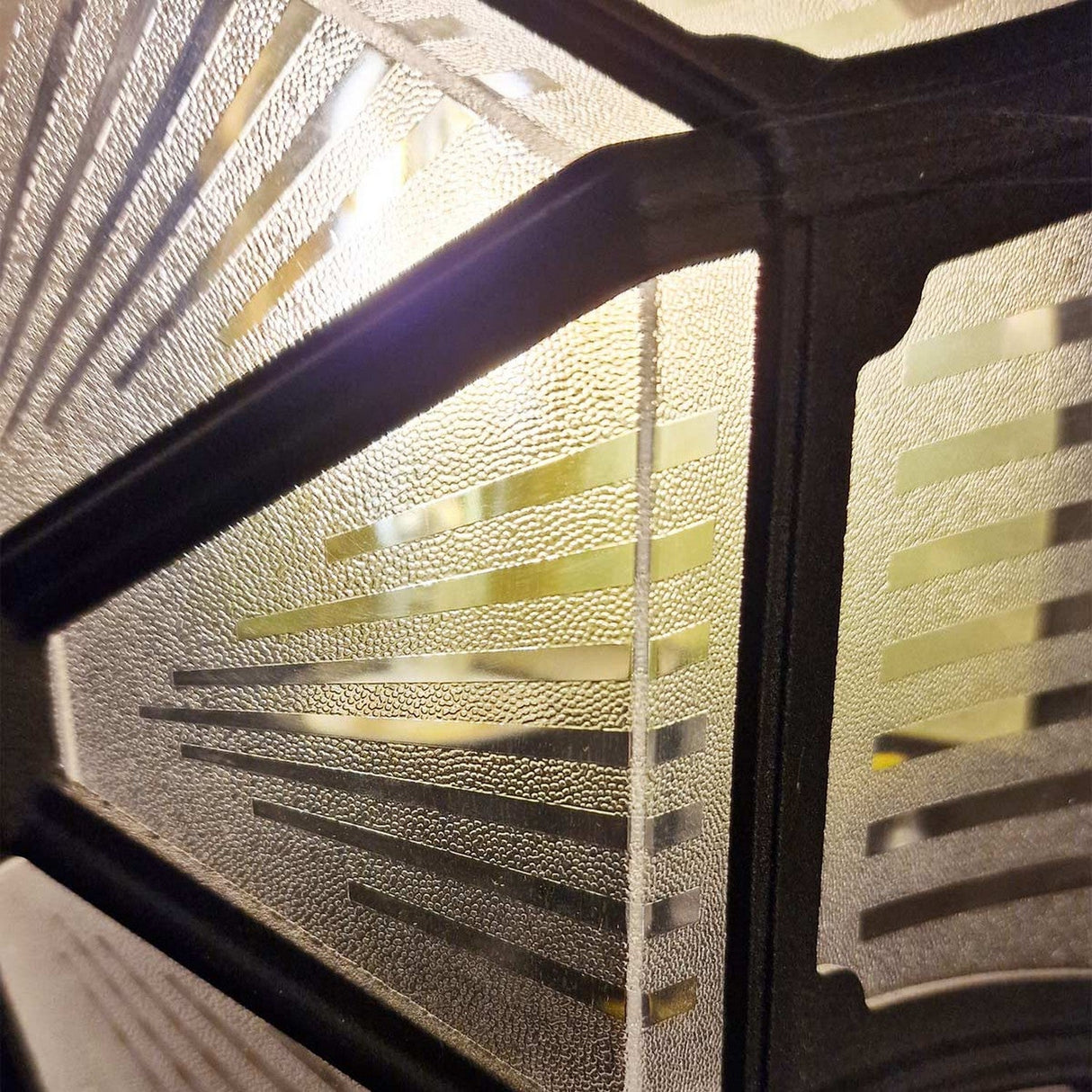 Close-up of the Parish 1 Light Outdoor Ceiling Flush - Black featuring a decorative glass panel with a geometric design. Framed by die-cast aluminum dividers, the pattern showcases angled lines that create a sunburst effect. Light filters through its IP44-rated textured glass surface, enhancing its intricate elegance and durability.