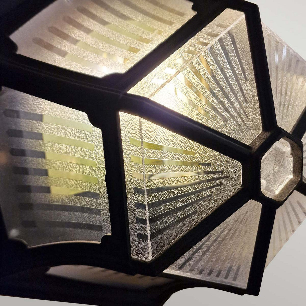 Close-up of the Parish 1 Light Outdoor Ceiling Flush - Black, featuring a geometric, star-like design. The frosted glass panels are framed by sturdy die-cast aluminum arranged symmetrically with etched radiating lines, creating a bright glow at the center. With an IP44 rating, it offers durability in various weather conditions.