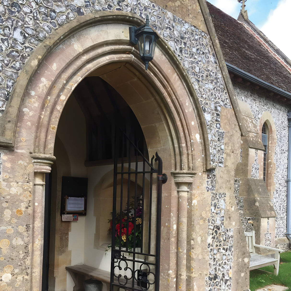 The stone church entrance, with its arched doorway and wrought iron gate, is graced by the Parish 1 Light Outdoor Down Wall Light in a black finish. Inside, vibrant flowers rest on a ledge. The exterior highlights textured stonework beneath a partly cloudy sky, accentuated by die-cast aluminum details.