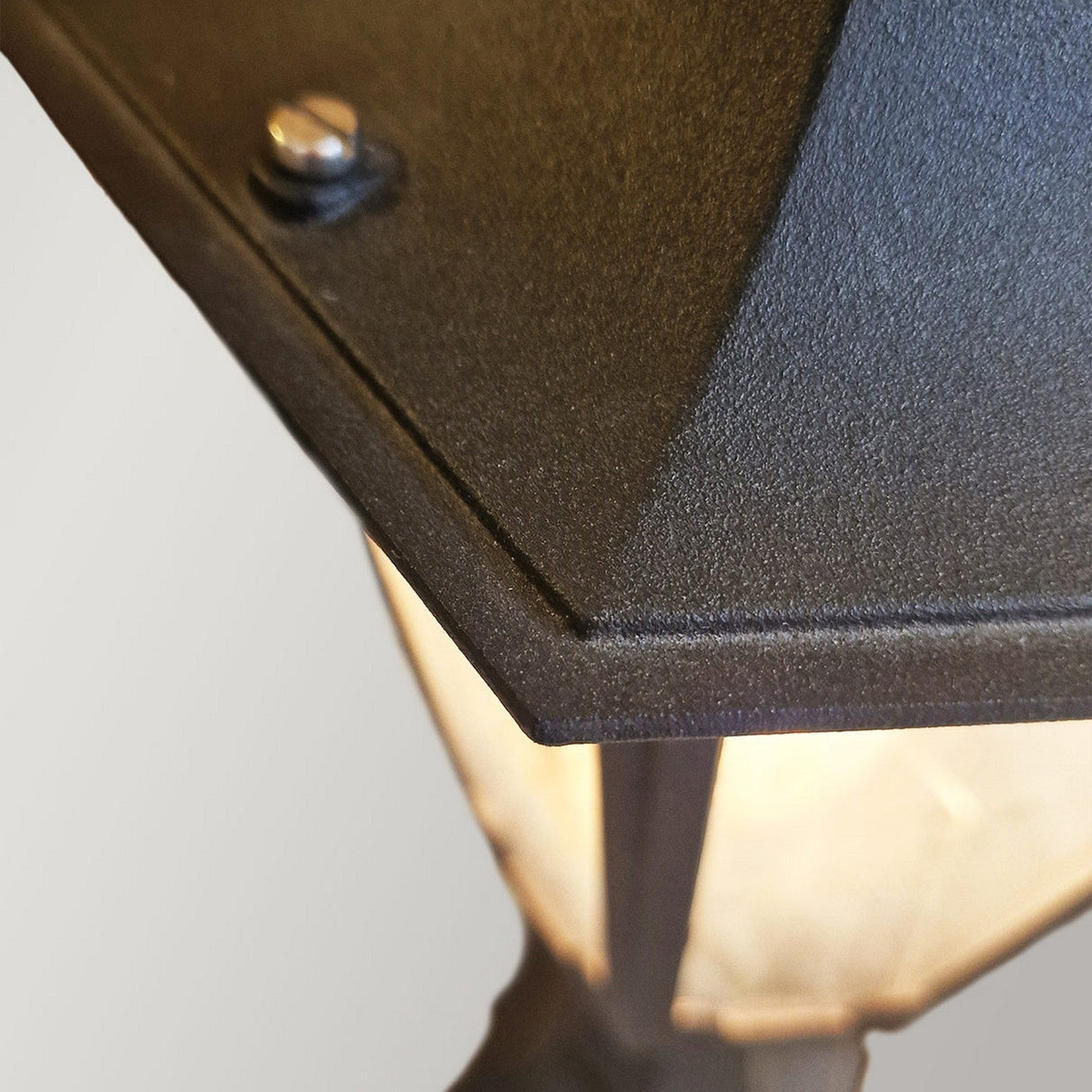 A close-up view of the Parish 1 Light Pedestal Light - Black reveals its triangular metallic structure with a visible bolt on the edge, partially covering a softly glowing light underneath. Crafted from die-cast aluminum, this lantern-style design elegantly contrasts against the plain, light gray background.