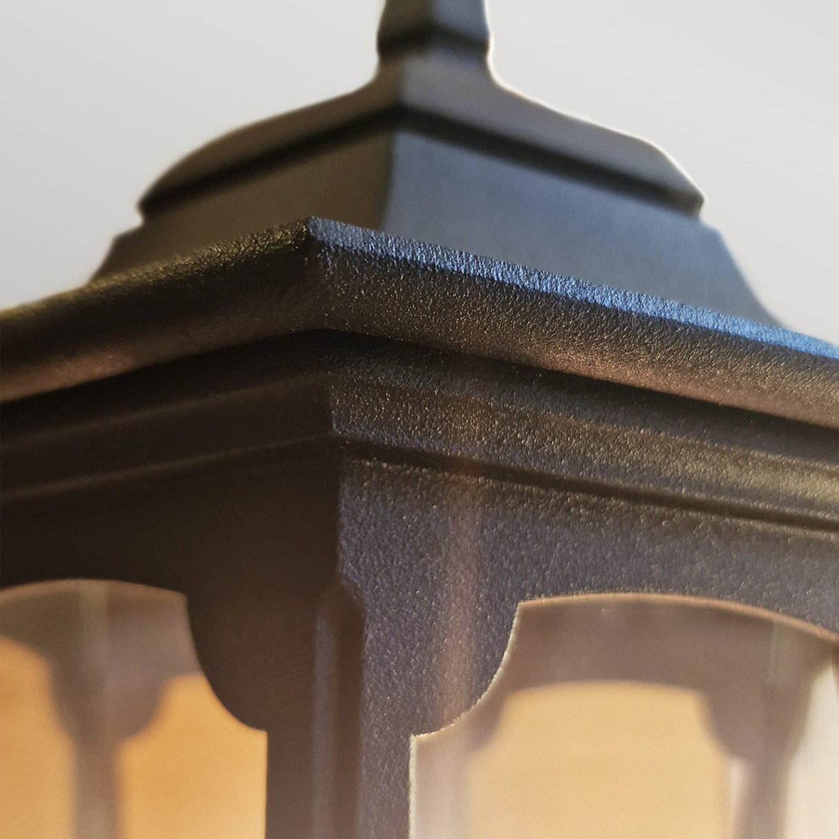 Close-up of the Parish 1 Light Pedestal Light - Black, featuring sharp edges and a textured surface crafted from die-cast aluminum. The prominent top showcases a classic lantern-style design against a softly blurred background.