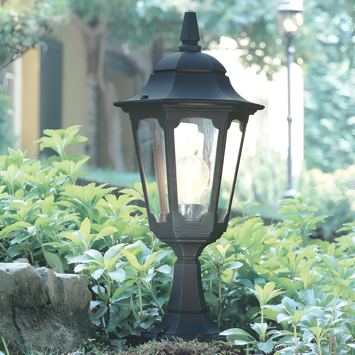 The Parish 1 Light Pedestal Light - Black, featuring a glowing bulb, stands amidst green foliage in a garden. Constructed from die-cast aluminium, its lantern-style design enhances the charm of the scene. A stone border is visible at the base, with blurred greenery forming the background.