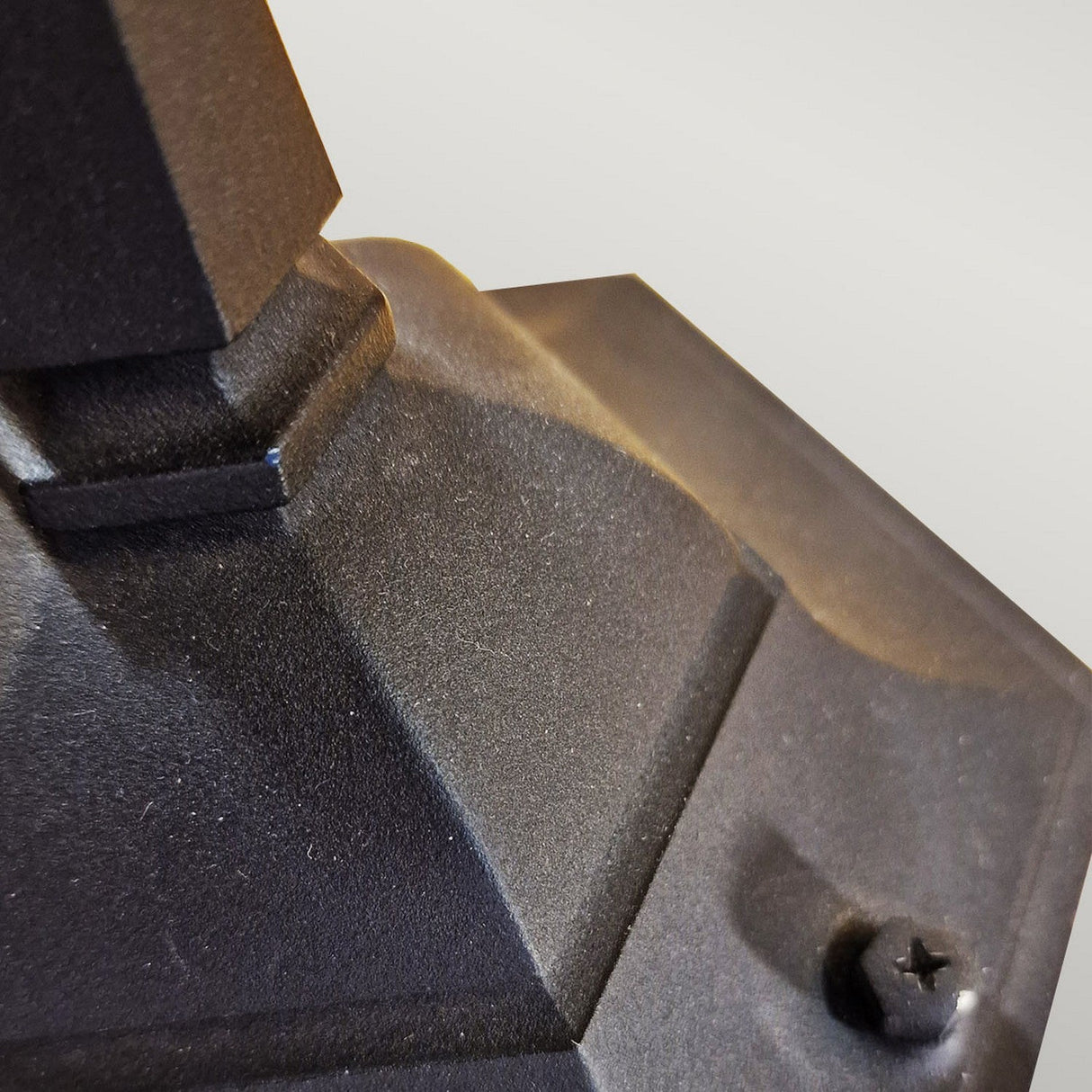 Close-up of the Parish 1 Light Pillar Light in black, featuring a dark, angular metal structure with a textured surface evocative of outdoor lighting designs. A six-pointed star-shaped screw is visible at the bottom right. The blurred background adds an element of mystery to this scene inspired by pillar lights.