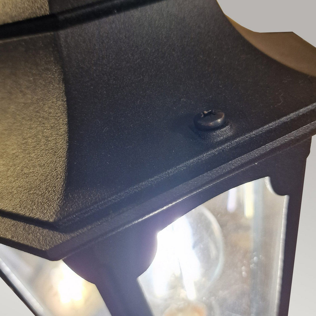 Close-up of the Parish 1 Light Lamp Post in black, crafted from die-cast aluminum and featuring a clear glass panel with a visible screw on top. The glowing light inside enhances its classic design with a slightly curved peak, making it an elegant choice for outdoor lighting.