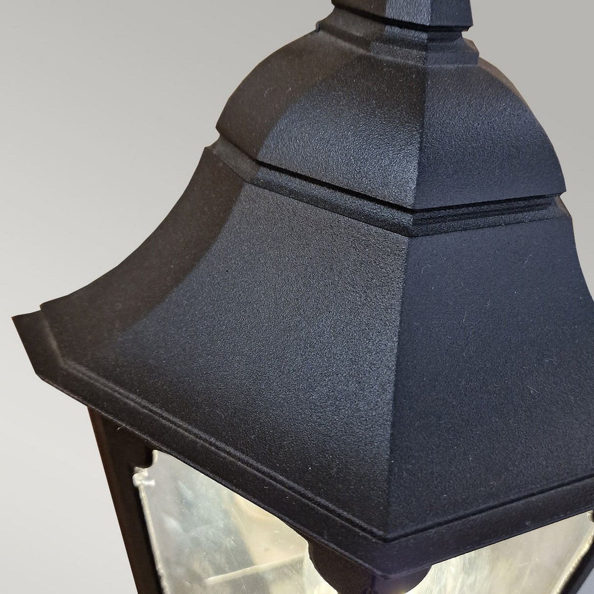 A close-up of the Parish 1 Light Outdoor Wall Light in black, featuring a pyramid-shaped top crafted from die-cast aluminum. The clear glass and plain gray background accentuate its design and texture, while its IP44 rating guarantees durability against the elements.