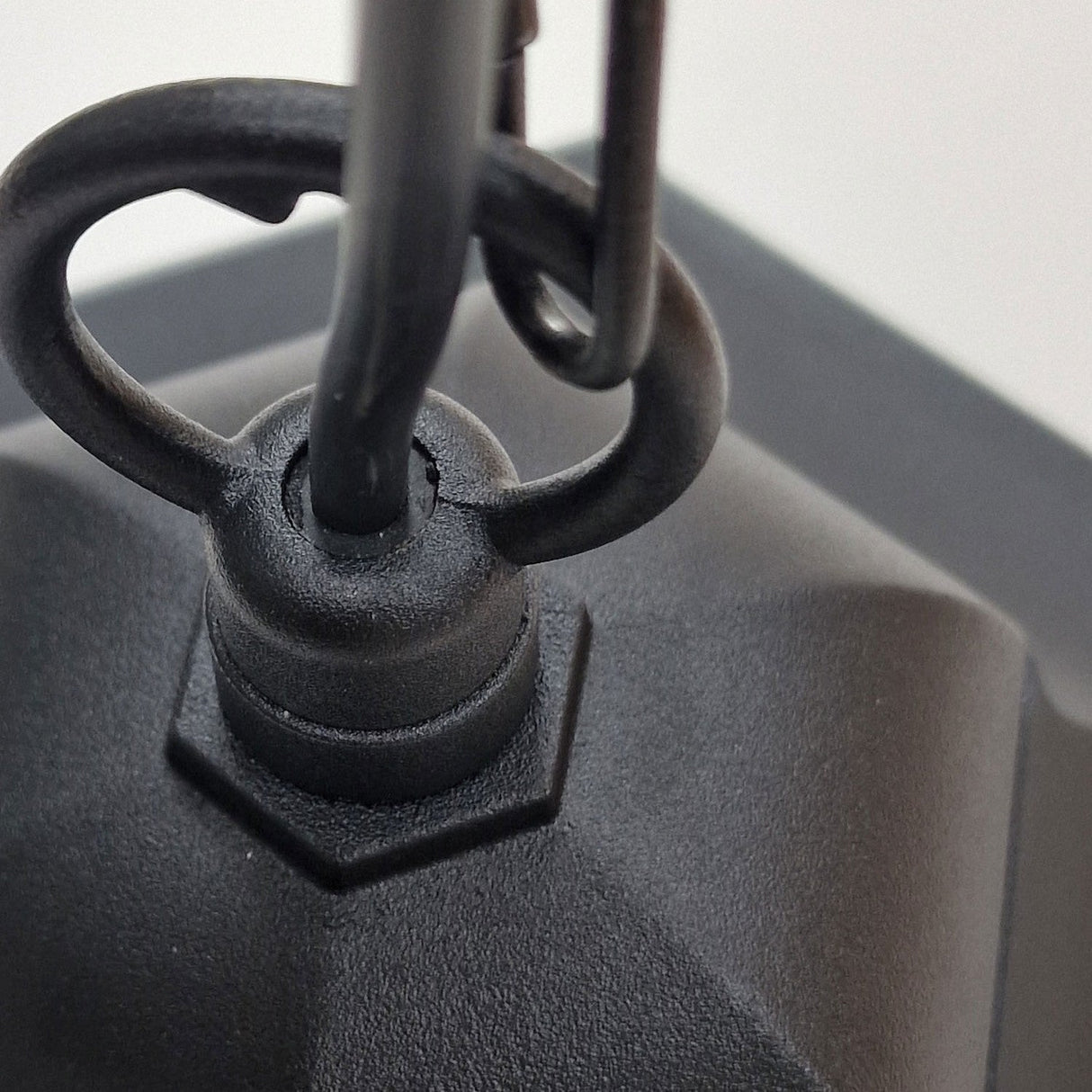 Close-up of the Parish 1 Light Outdoor Chain Pendant - Black, featuring a hexagonal metal knob with an attached loop, connected to a cylindrical rod. Crafted from premium die-cast aluminium, it showcases a matte black finish. The slightly blurred background accentuates its outdoor chain pendant design, infusing vintage charm into any setting.