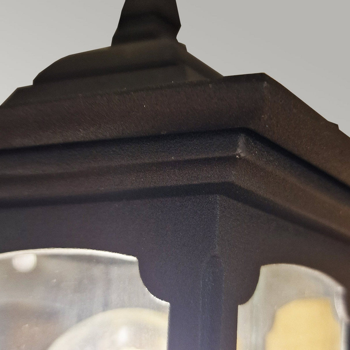 Close-up of the Parish Mini 1 Light Outdoor Up Wall Light in black, featuring glass panels that highlight the textured die-cast aluminum frame and a visible light bulb inside. The background displays a soft gradient of gray.