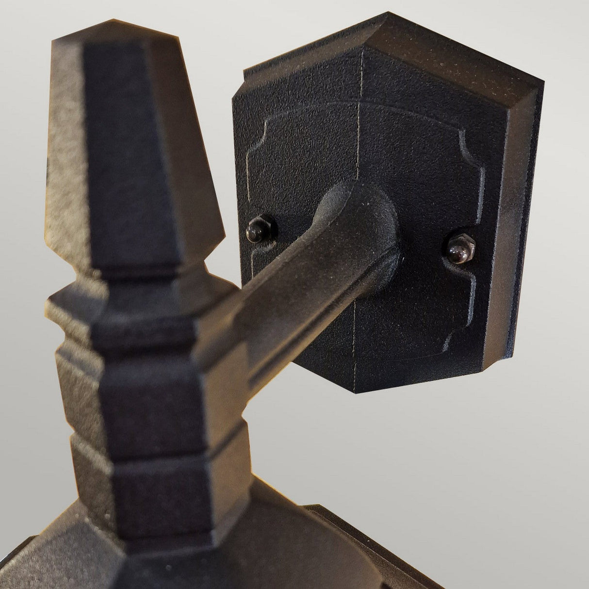 Close-up view of the Parish Mini 1 Outdoor Light Down Wall Light in black. This wall-mounted fixture features geometric designs and a textured finish, complete with a sturdy arm and decorative top. Perfect for illuminating exterior walls, it is set against a neutral light gray background.