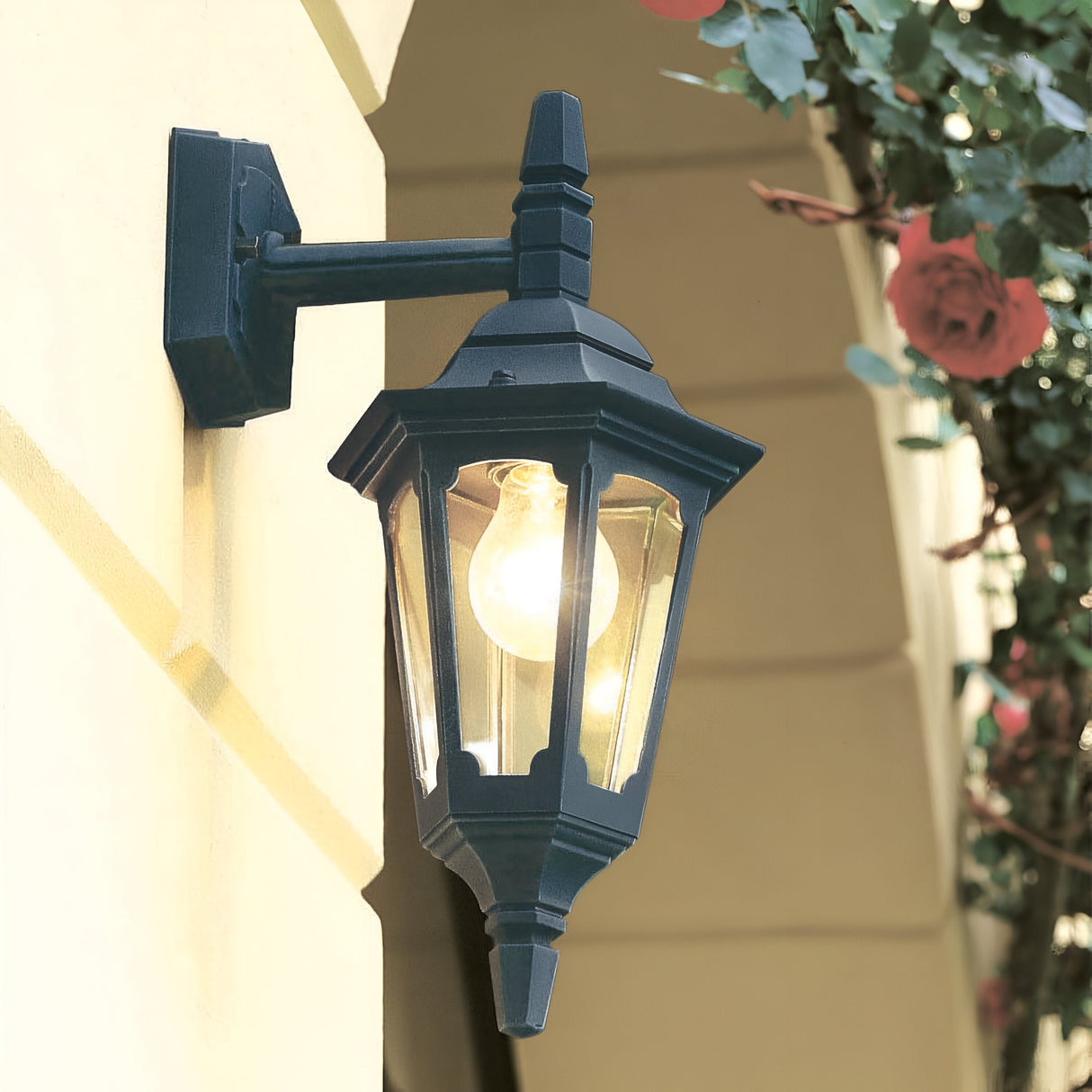 The Parish Mini 1 Outdoor Light Down Wall Light in Black is affixed to a light-colored exterior wall, with its illuminated bulb casting a warm glow. This down-facing light enhances the greenery and pink flowers above on the right side, establishing an inviting ambiance.