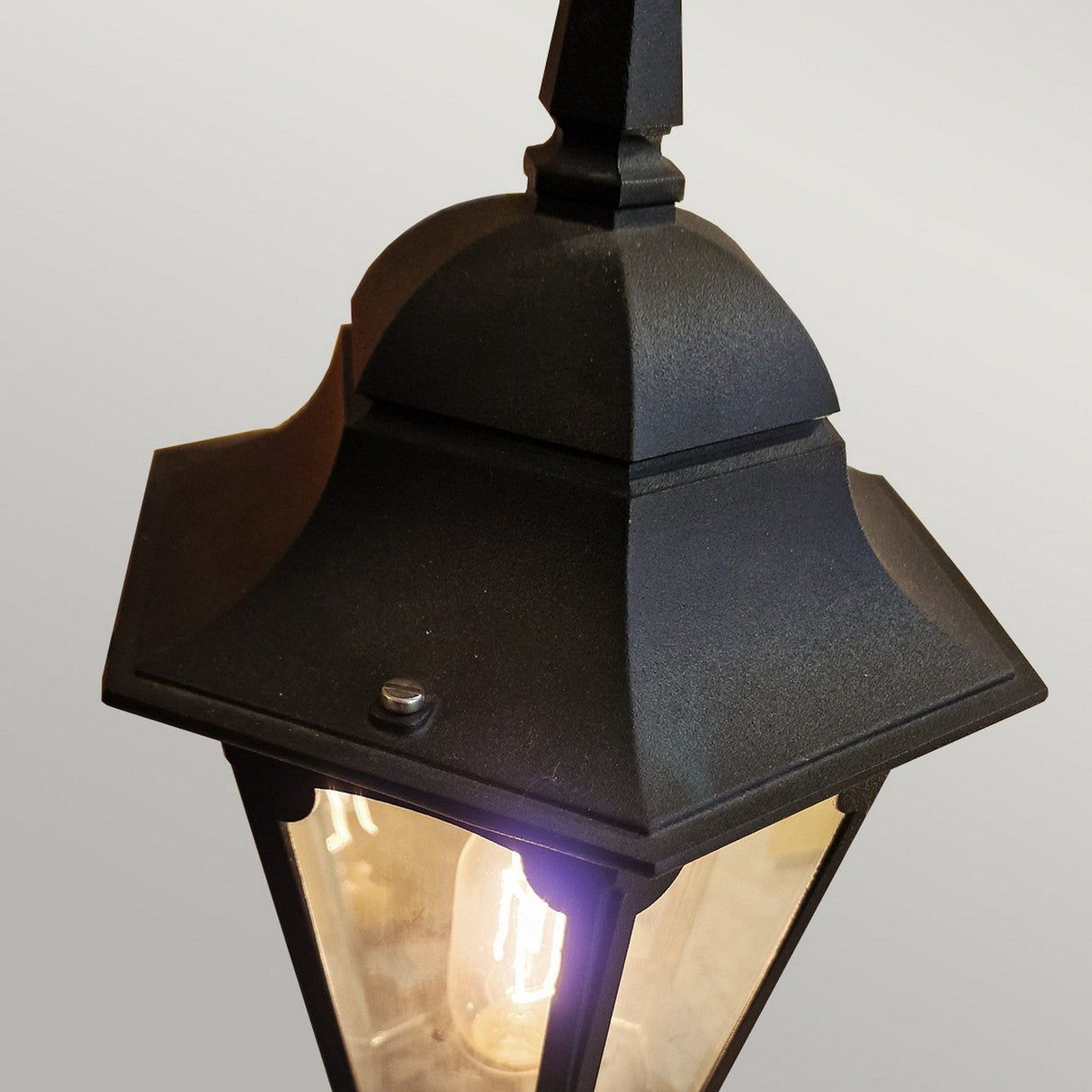 A close-up view of the Parish Mini 1 Light Pedestal Light - Black, featuring a vintage design with clear glass that highlights a glowing bulb. This classic outdoor lantern is mounted on a wall, enhancing the ambiance against a neutral gray background.