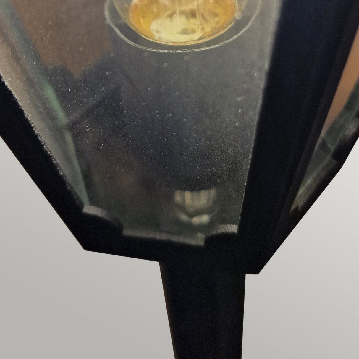 Close-up view of the Parish Mini 1 Light Pillar in Black, featuring a glowing bulb inside and showcasing a traditional lantern design. The frosted glass panels and black die-cast aluminum frame create a classic look, set against a muted gray background.