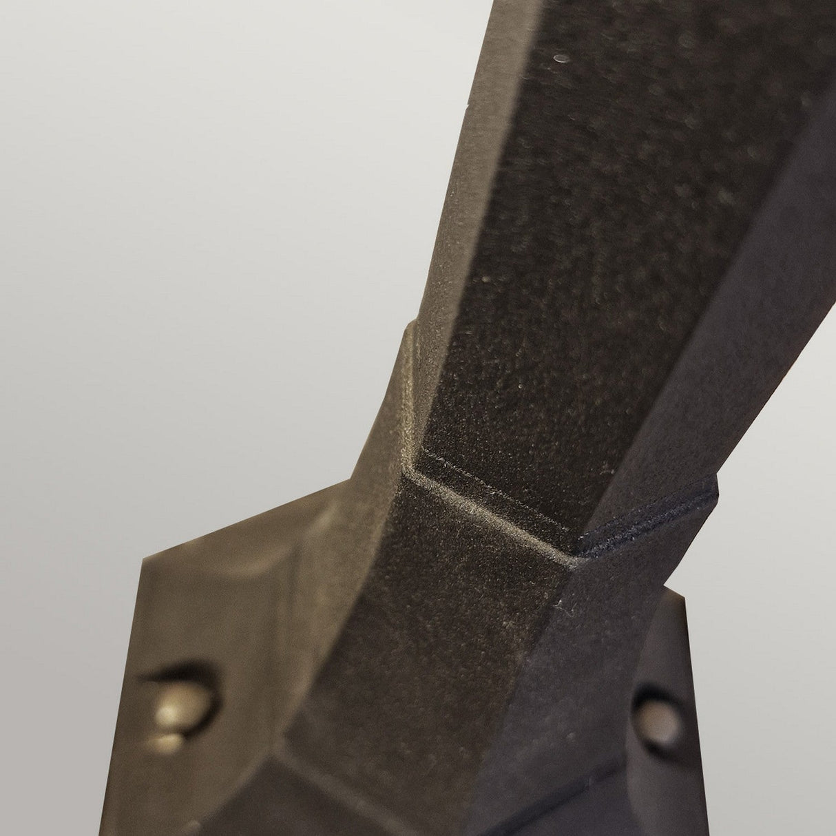 The image showcases the Parish Mini 1 Light Pillar - Black, highlighting its textured, angular design crafted from die-cast aluminum. It's mounted on a square base against a plain, light gray backdrop.