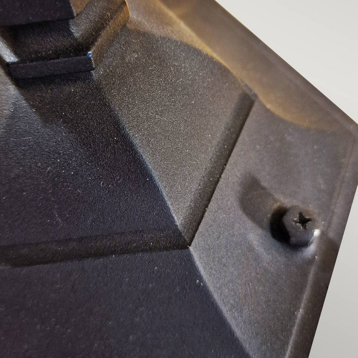 Close-up view of the Parish Mini 1 Light Pillar - Black, showcasing its textured, dark surface with geometric patterns crafted from die-cast aluminum. A small, star-shaped bolt sits on the right side, with the angle and lighting creating a sense of depth and shadow reminiscent of traditional lantern design.