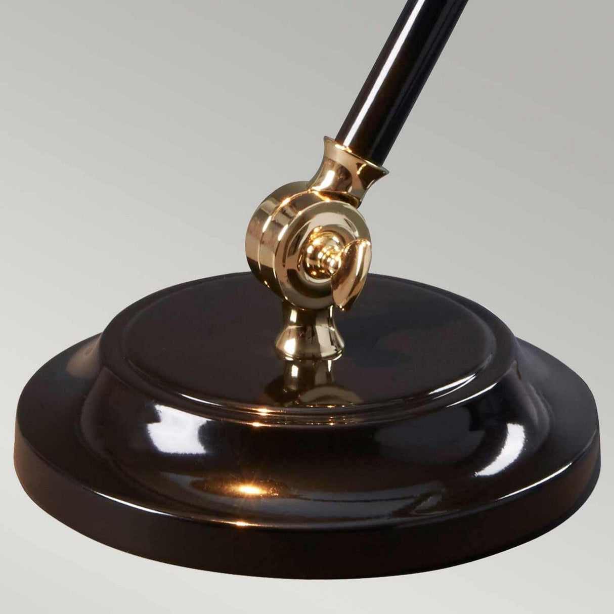 Close-up of the round, polished brass base of the Provence Element 1 Light Mini Table Lamp - Black/Polished Brass, featuring a gold-colored adjustable joint. The lamp's support extends upward, highlighting its French-inspired design against a smooth gradient gray background.