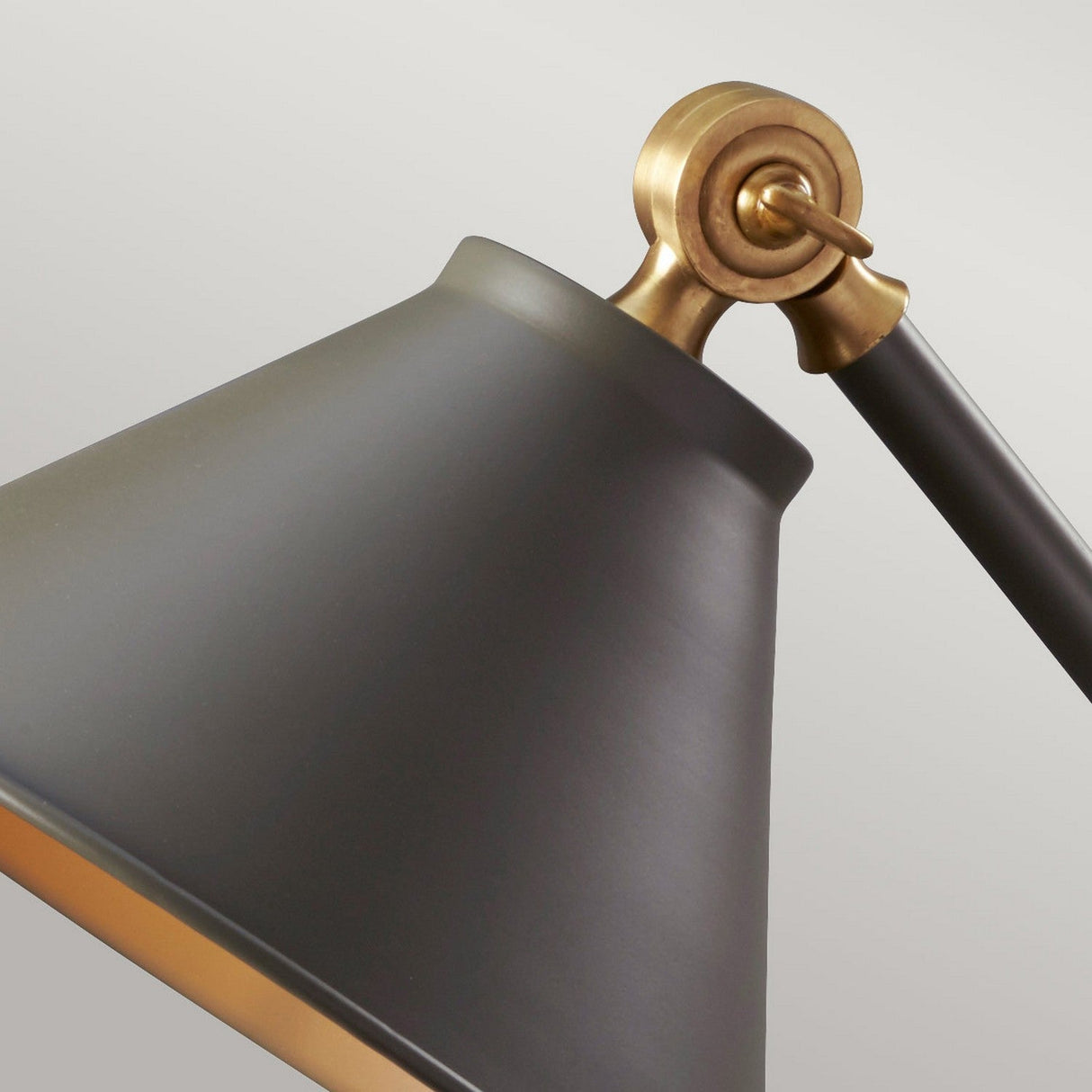 A close-up of the Provence Element 1 Light Mini Table Lamp in dark grey with aged brass accents shows off its matte conical shade against a light gray background. This piece elegantly combines vintage and modern design elements and features an adjustable angle with its pivot joint.