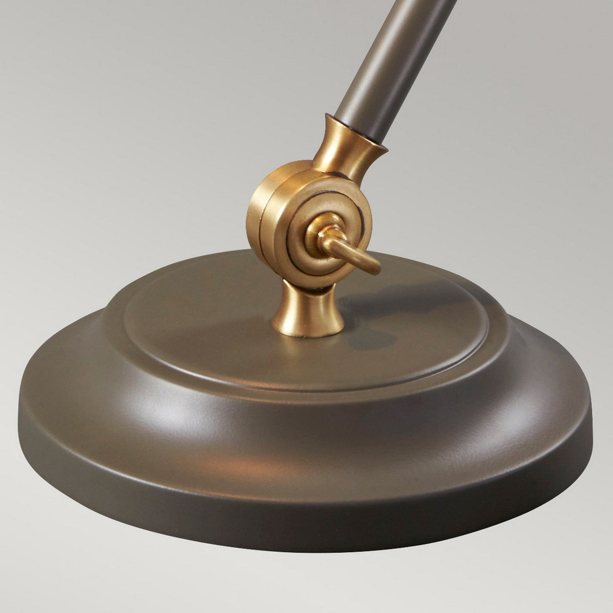 A close-up of the Provence Element 1 Light Mini Table Lamp showcases its dark grey, round base and an aged brass joint with a screw, where vintage meets modern. The design highlights the lamp's adjustable feature against a neutral light gray background.