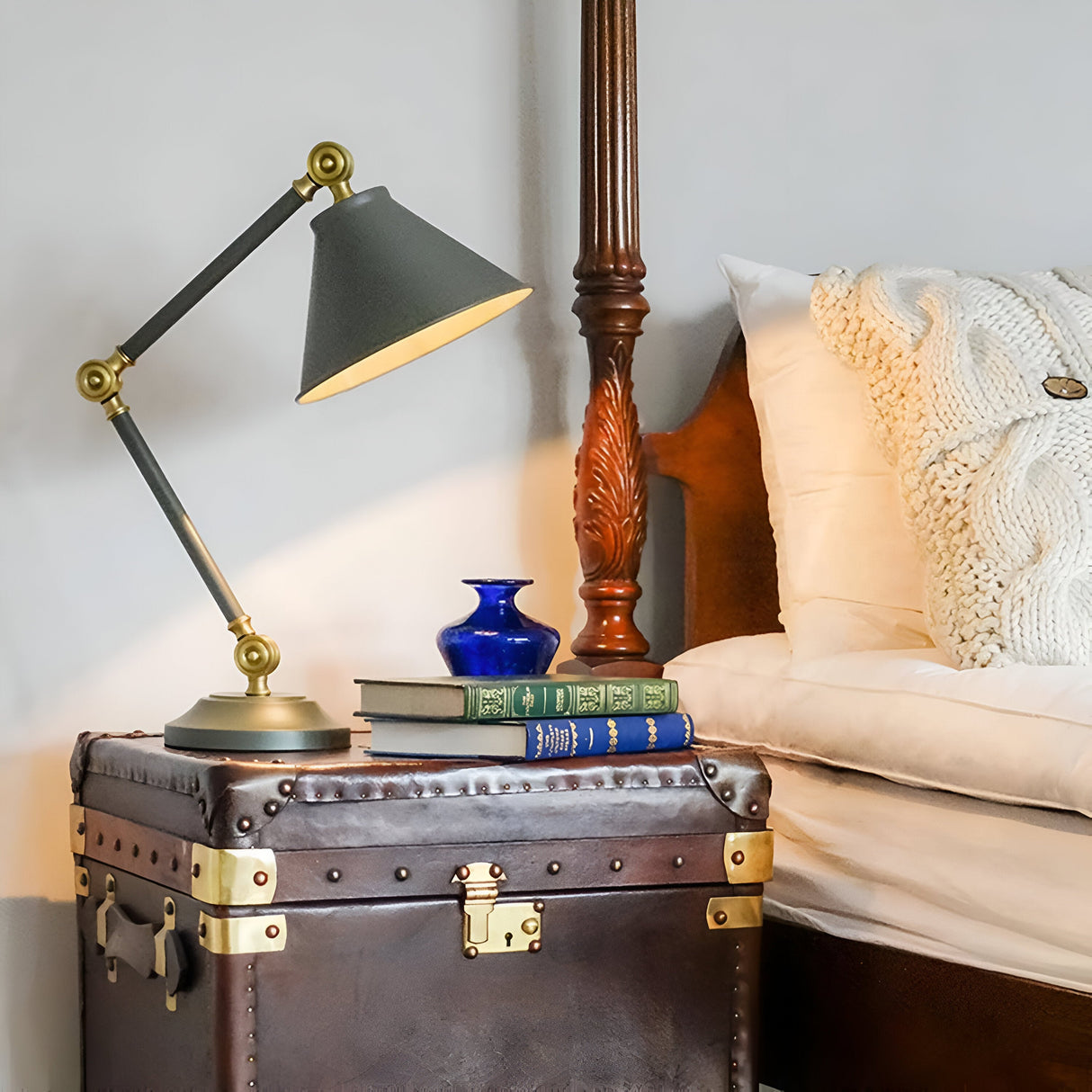 A Provence Element 1 Light Mini Table Lamp in dark grey with aged brass accents is mounted on a brown leather trunk featuring aged brass highlights. Next to it, a small blue vase and several books enhance the charm. This trunk, which seamlessly combines vintage and modern design elements, functions as a nightstand beside a bed adorned with a white knitted blanket.