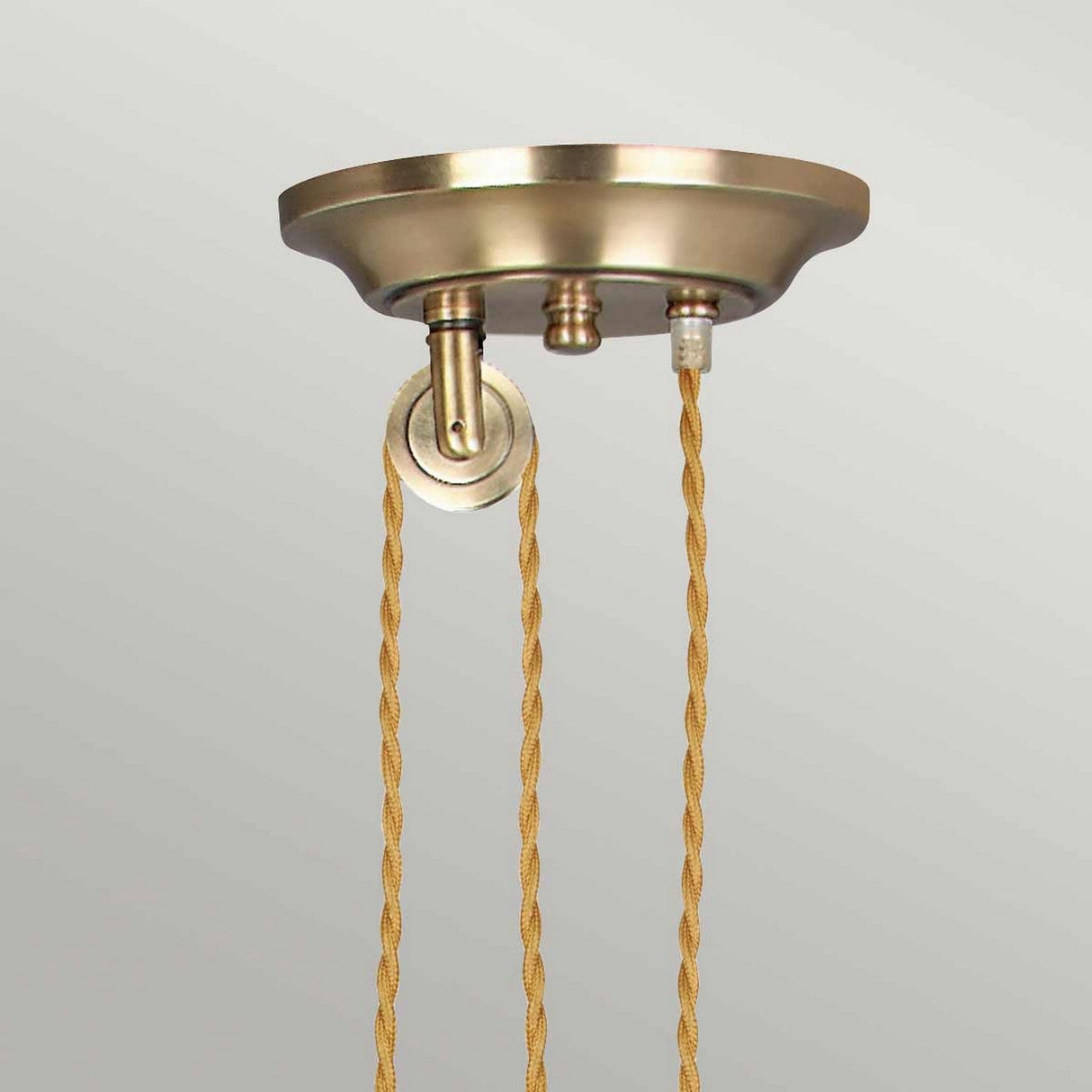 A detailed view of the Provence 1 Light Rise & Fall Pendant in aged brass, showcasing two gracefully twisting gold-colored cords descending from the vintage-inspired ceiling canopy. These cords are attached to the canopy through distinct fixtures, set against a simple light gray backdrop.