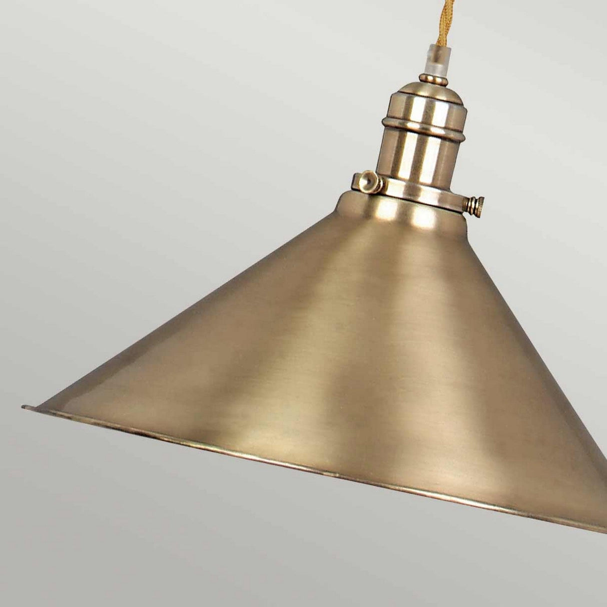 The Provence 1 Light Rise & Fall Pendant in aged brass, showcasing a conical shade and metallic industrial design, is suspended by a cord against a plain gray background, exuding a sleek and modern aesthetic.