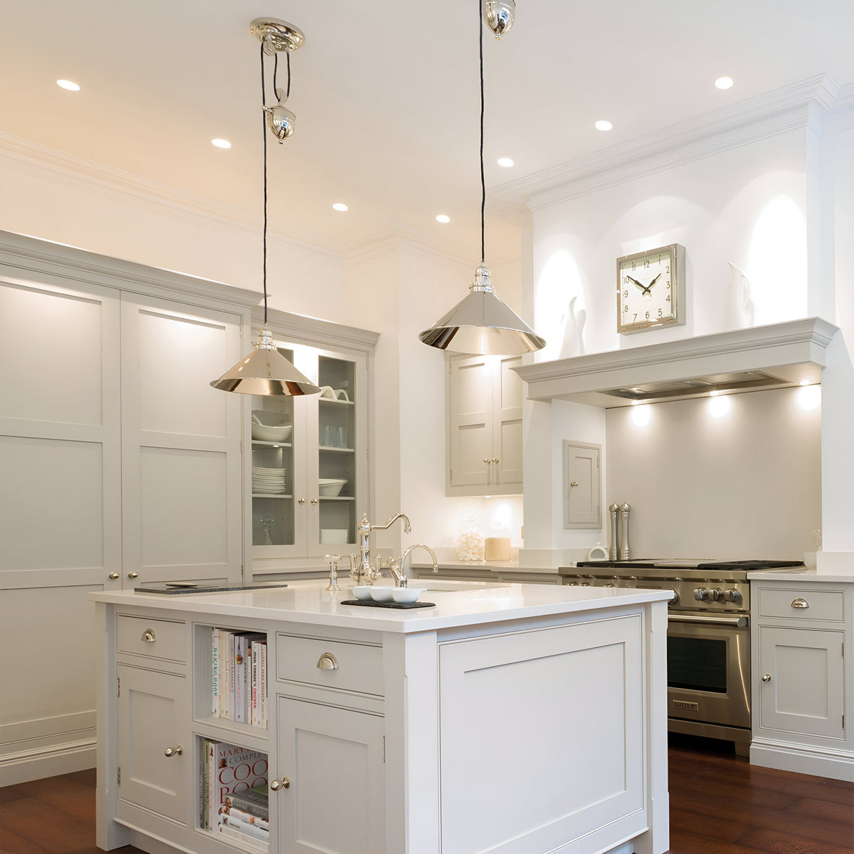 A modern kitchen features white cabinets and a large island with built-in bookshelves. Two Provence 1 Light Rise & Fall Pendants in polished nickel hang above the island, while stainless steel appliances, including an oven and stove, are positioned against the back wall with a clock mounted above the stove.