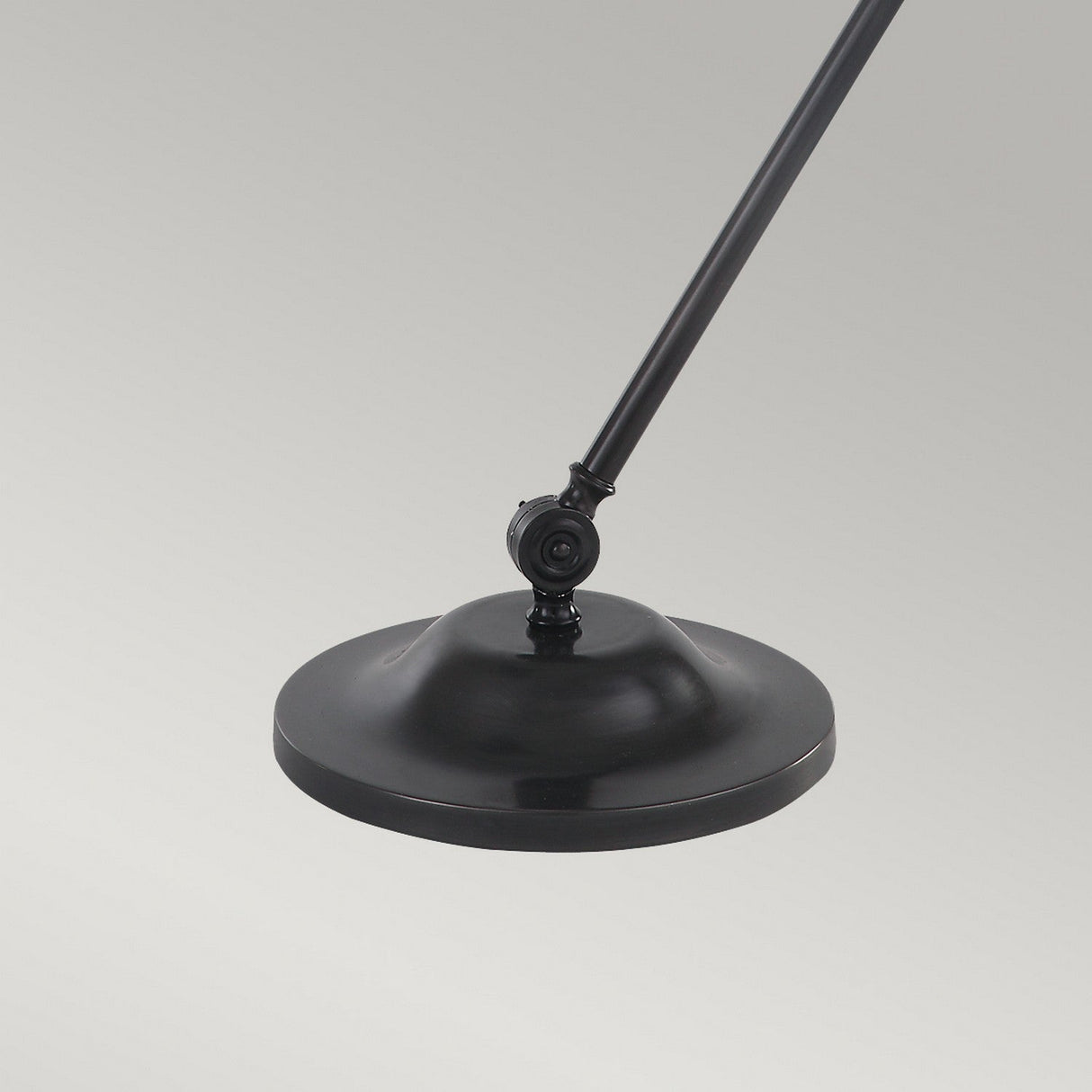 Close-up of the round base of the Provence 1 Light Table Lamp in Old Bronze, featuring a sleek, slender arm extending upward. Its sophisticated design includes adjustable knuckle joints that create a striking contrast against the plain gray backdrop.