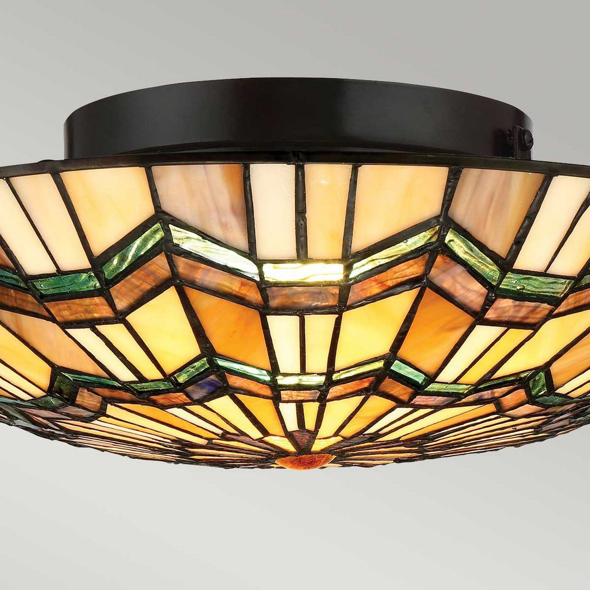 The Alcott 2 Light Tiffany Flush in Valiant Bronze features a stained glass ceiling design showcasing geometric patterns in shades of yellow, orange, and green. Drawing inspiration from the Arts and Crafts movement, it emits a warm, diffused glow against a neutral backdrop.