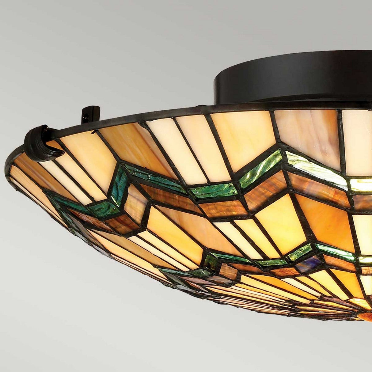 A close-up of the Alcott 2 Light Tiffany Flush in Valiant Bronze showcases geometric patterns in shades of amber, cream, and green. The light is mounted on a dark fixture, casting a warm glow reminiscent of the Arts and Crafts movement.