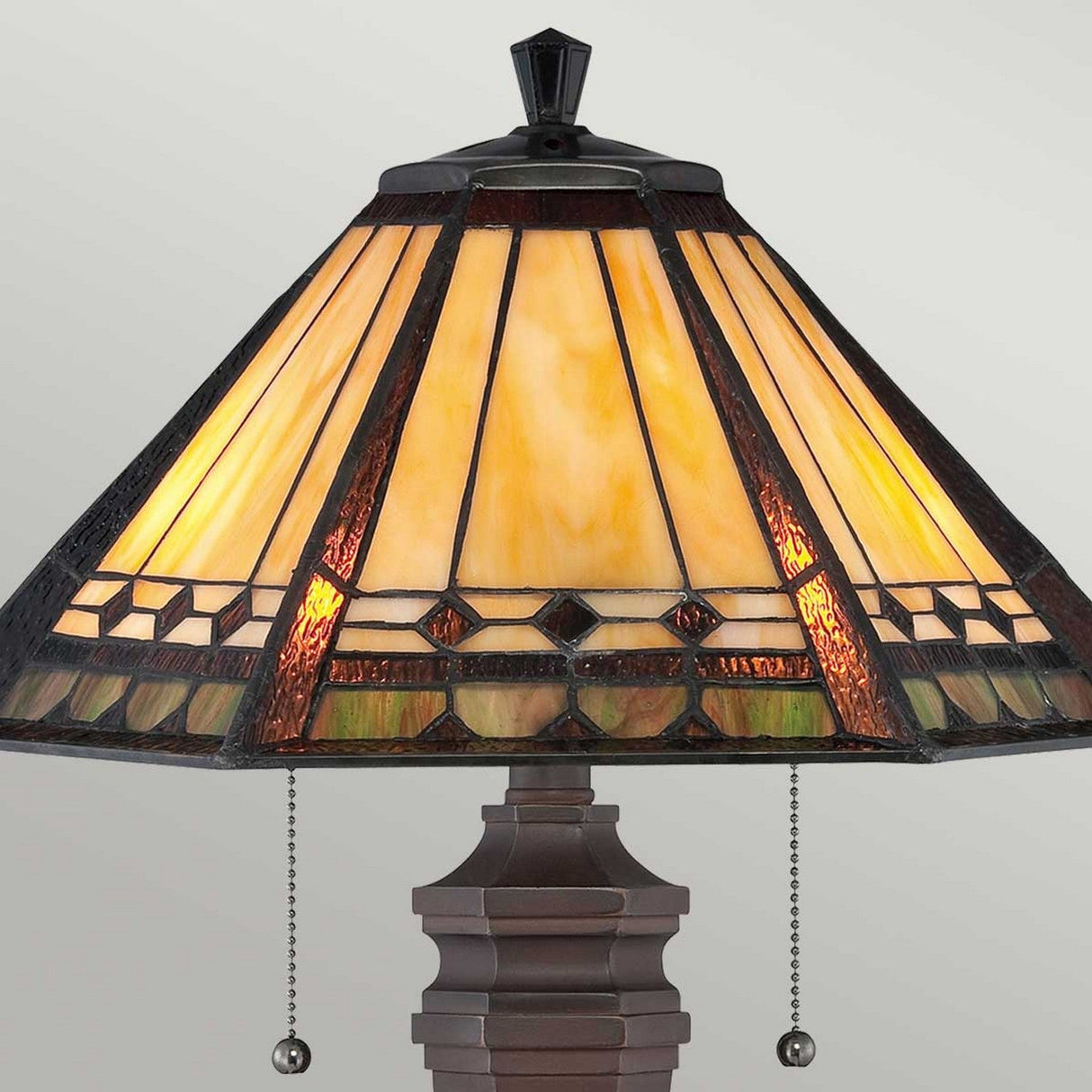 The Arden 2 Light Table Lamp - Bronze Patina features a pyramid-shaped stained glass shade with geometric patterns in warm amber and brown tones. The pull chains are embellished with decorative beads, and the lamp's bronze patina finish enhances its elegance. When illuminated, it emits a soft, inviting glow.