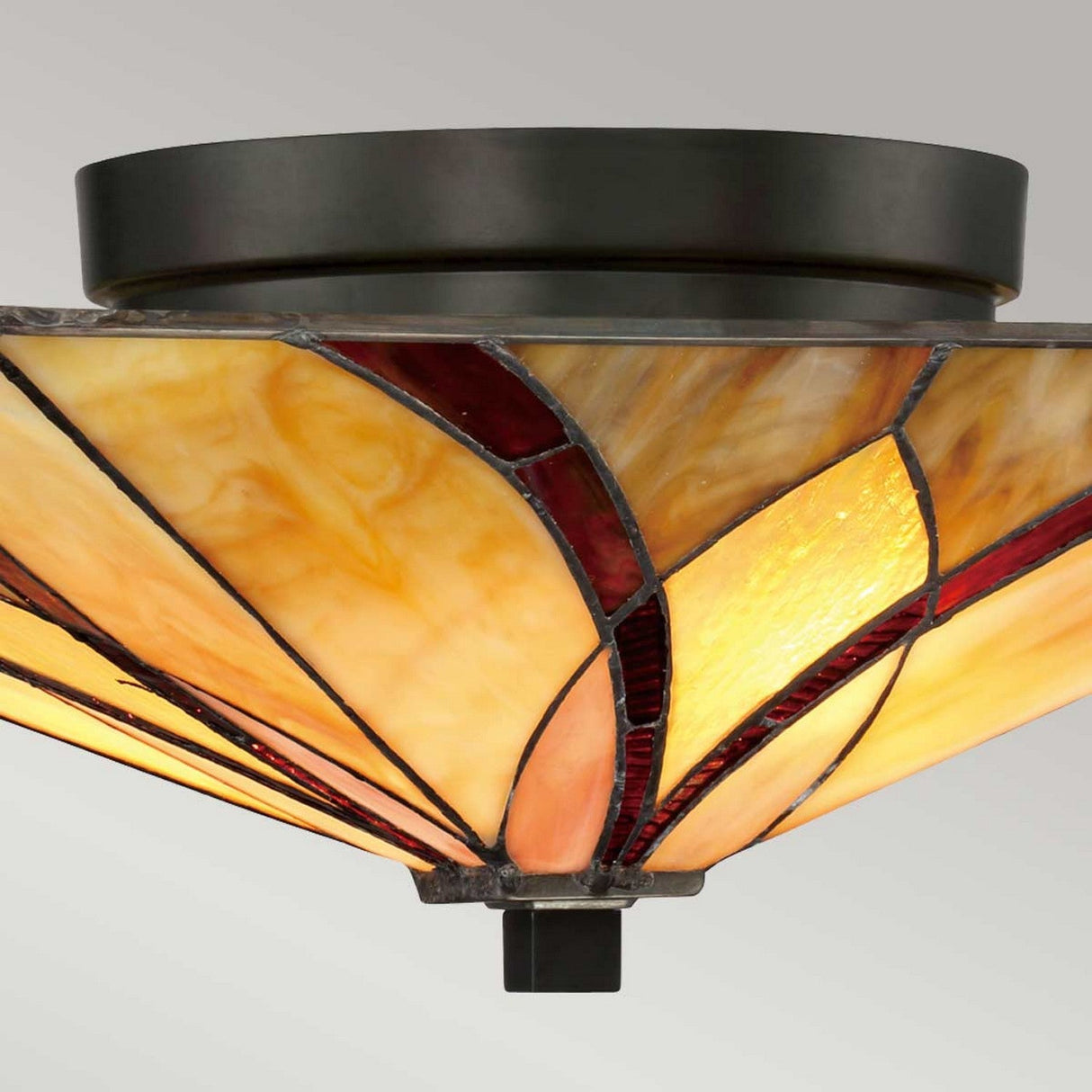 A close-up view of the Asheville 2 Light Tiffany Flush in Valiant Bronze reveals a stained glass ceiling light fixture featuring a geometric design with warm amber and burgundy panels, reminiscent of a classic Tiffany style.