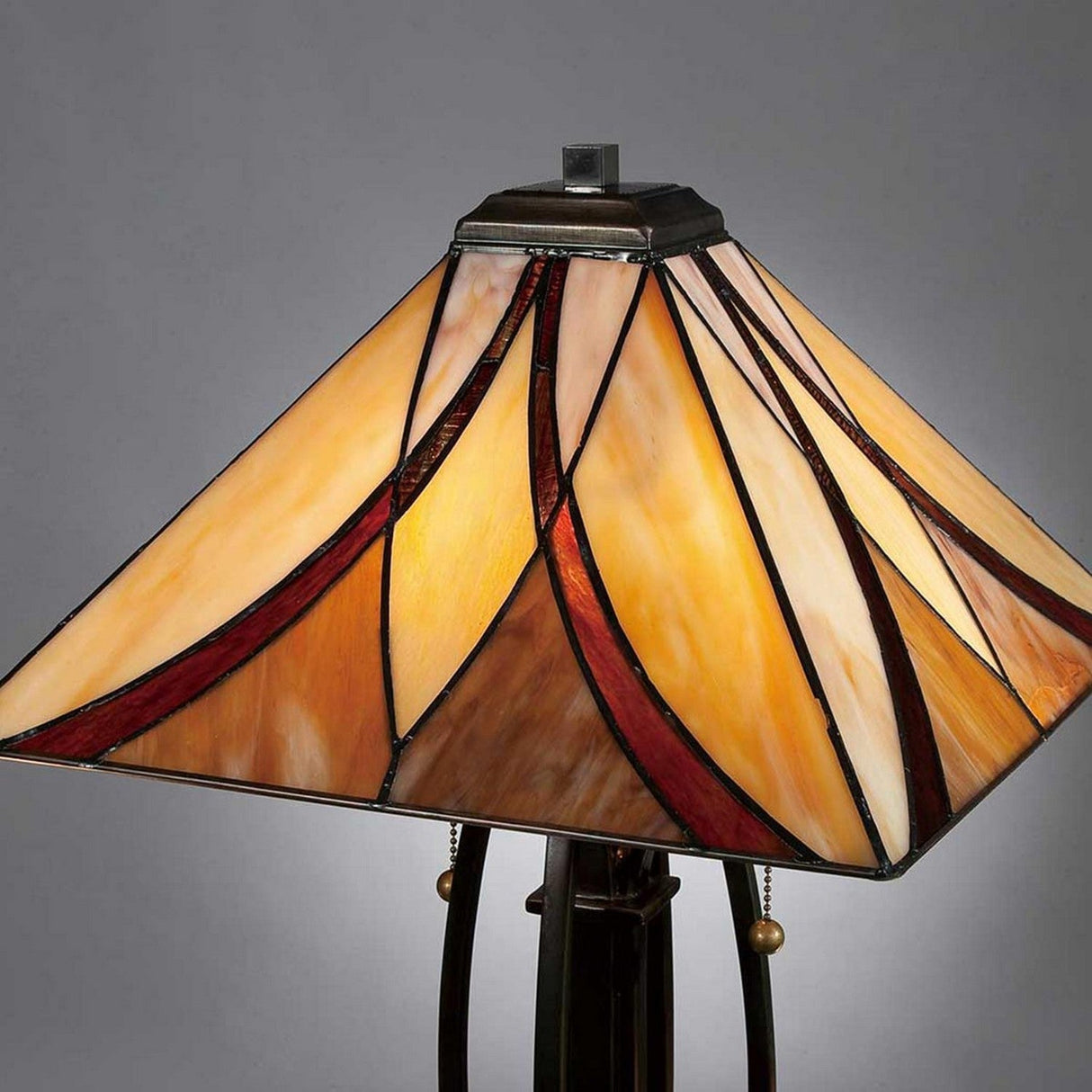 The Asheville 2 Light Tiffany Table Lamp - Valiant Bronze features a classic Tiffany-style shade in warm hues of orange, yellow, and red that sits elegantly against a plain gray background, with its base finished in Valiant Bronze.