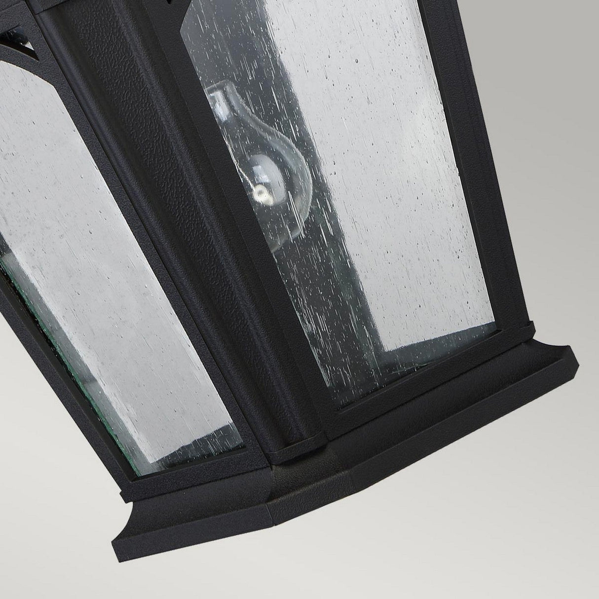 A close-up view of the Bedford Outdoor 1 Light Large Wall Lantern in black highlights its clear glass pane, dotted with raindrops. The visible light bulb inside is framed against a neutral gray background.