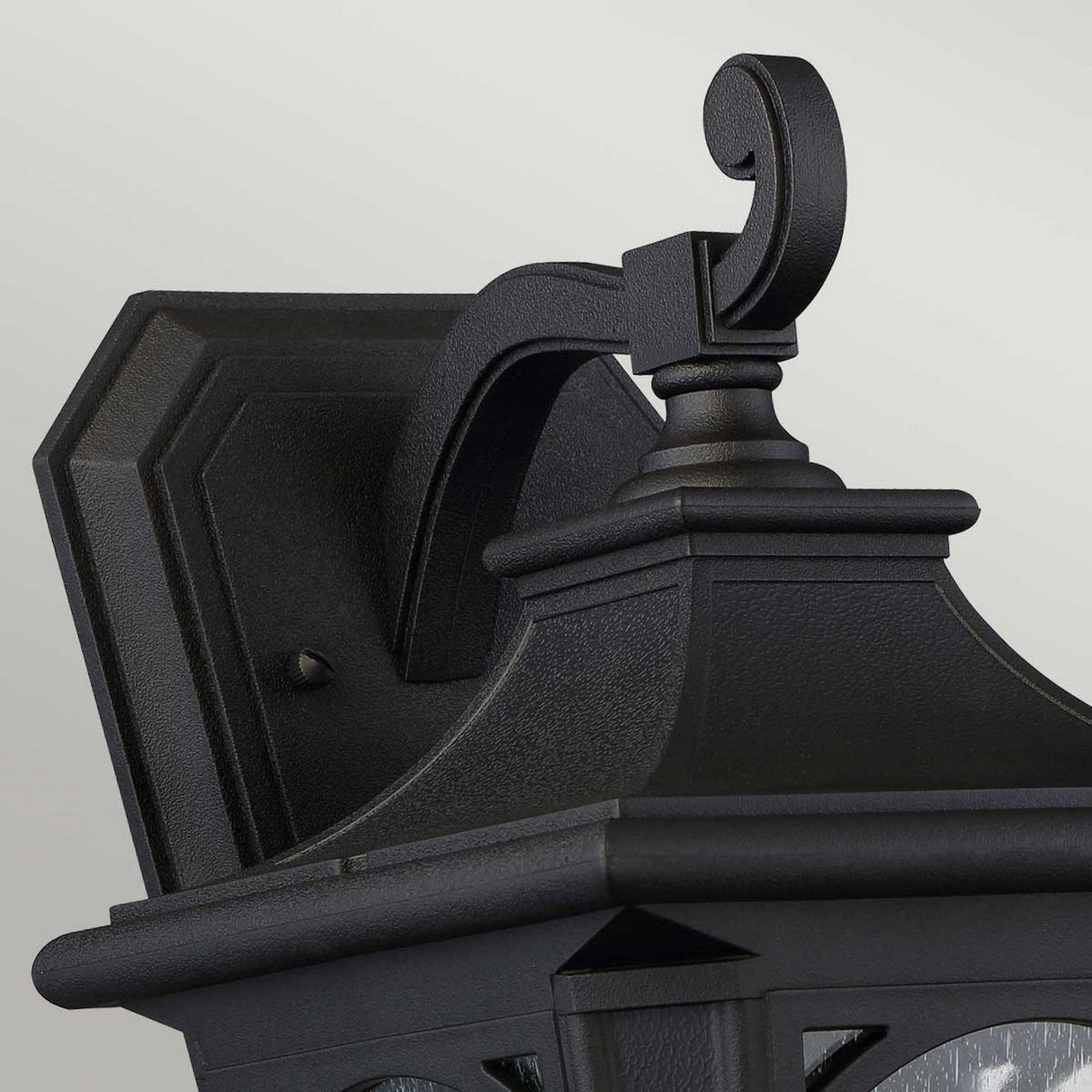 Close-up of the Bedford Outdoor 1 Light Medium Wall Lantern in Black, highlighting its decorative top and geometric base. Mounted on a bracket with a scrolling design, it features clear seedy glass panels against a plain gray wall, providing durable outdoor lighting.