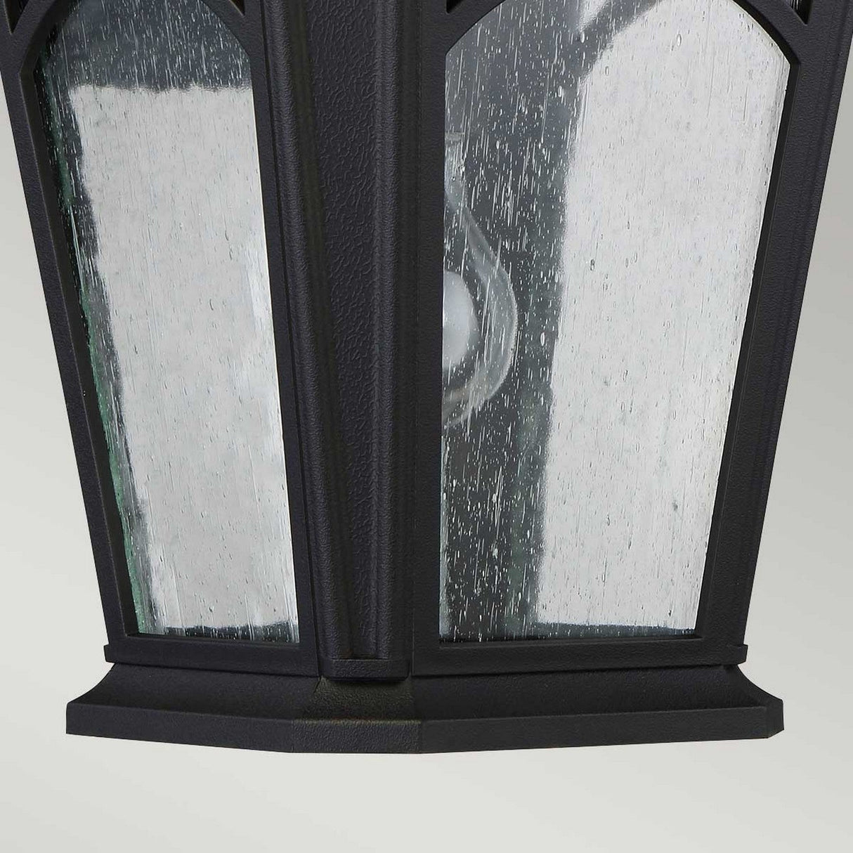 Close-up of the Bedford Outdoor 1 Light Medium Wall Lantern - Black, showcasing its durable outdoor lighting. The glass panel features a textured, raindrop-like finish similar to clear seedy glass. Inside, a single light bulb provides a vintage-style design against a simple, light gray background.
